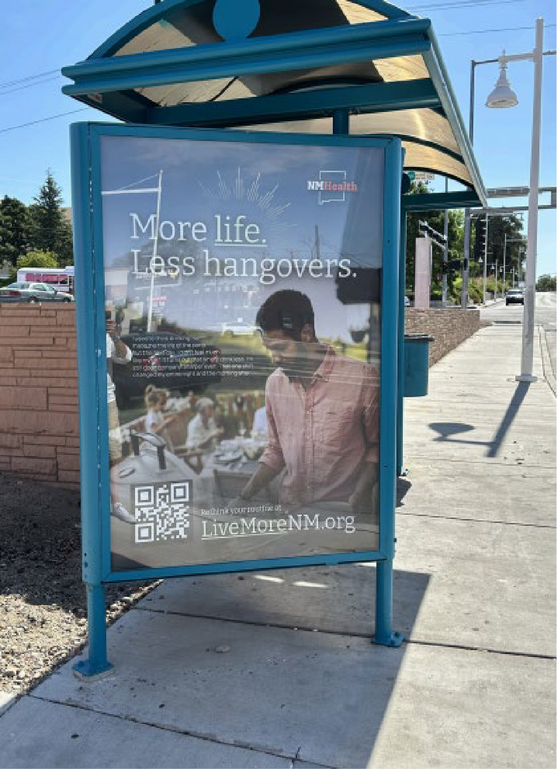 A bus stop sign with an advertisement that reads 'More life. Less hangovers.' It features a woman preparing food outdoors, with a QR code and website URL LiveMoreNM.org at the bottom.