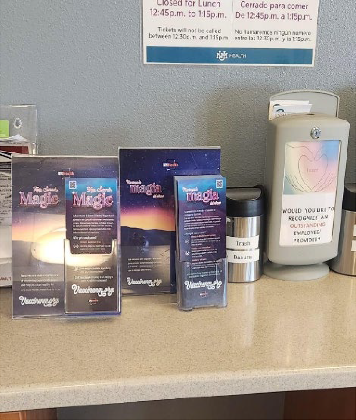 Brochures titled "Your Claims Made Easy" and "Tailored Imaging" displayed on a countertop near a trash can and a dispenser with a sign asking, "Would you like to recognize an outstanding employee?" A sign on the wall indicates lunch hours and ticket sale times.