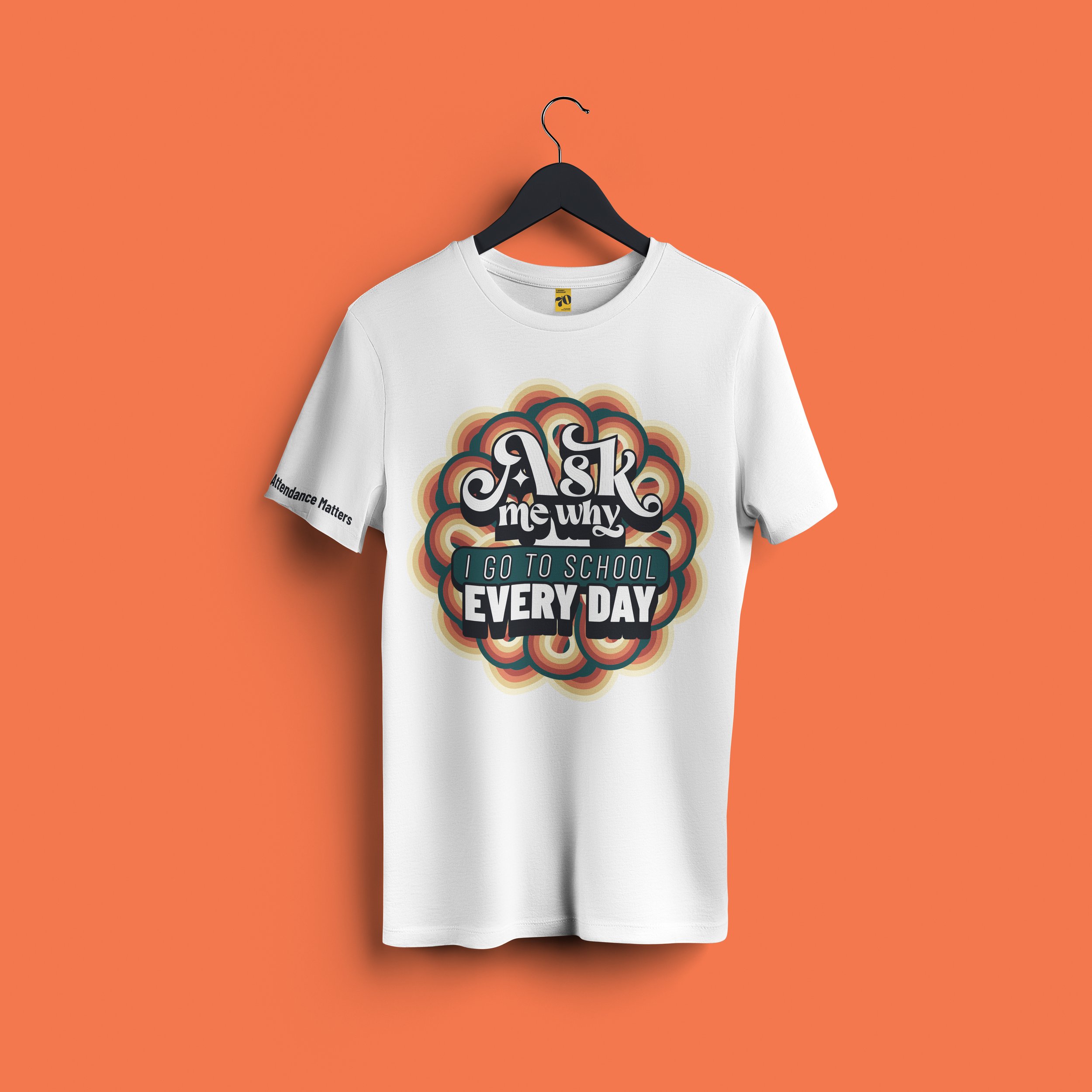 A white T-shirt on a black hanger against an orange background. The T-shirt has a colorful graphic design with the phrase "Ask me why I go to school every day" and a small black text on the sleeve that says "Attendance Matters".