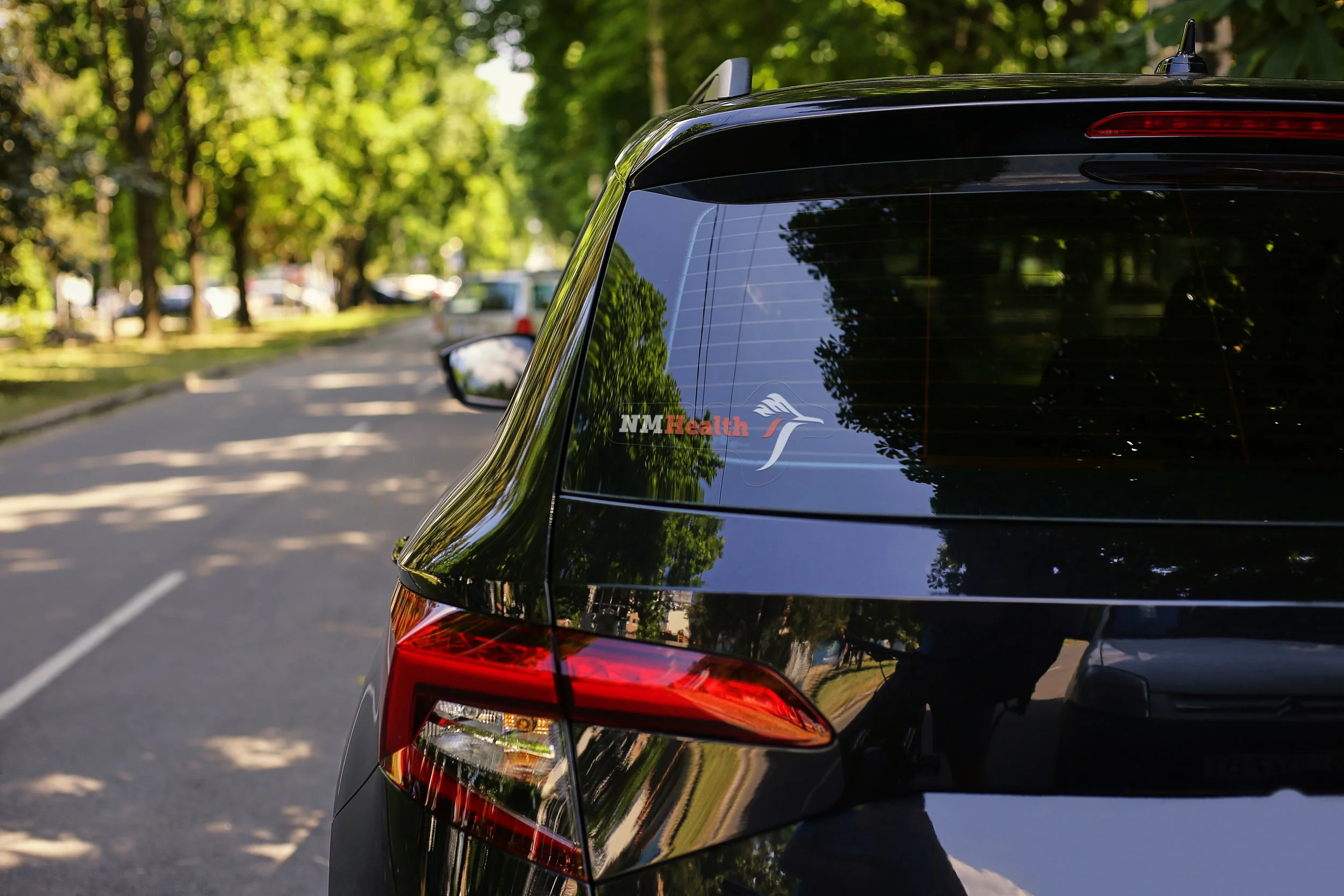 Close-up of the rear side of a black car parked on a street with trees and other vehicles in the background, featuring a sticker that reads 'NMHealth' with a stylized bird logo.