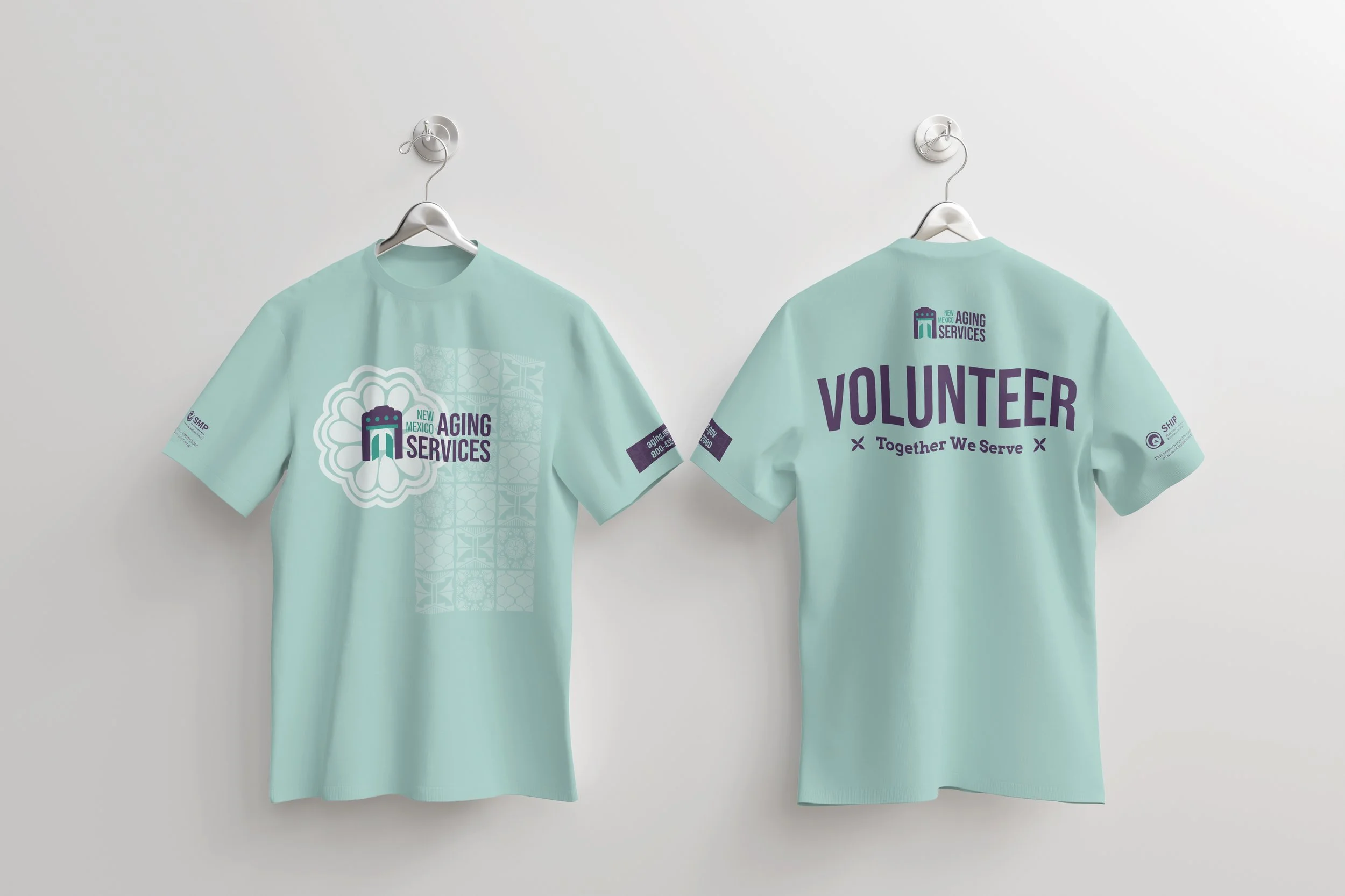 Two light blue T-shirts hanging on hooks against a white wall. The front of the shirts features a logo for New Mexico Aging Services, and the back has the word "VOLUNTEER" in large purple letters with the phrase "Together We Serve" underneath.