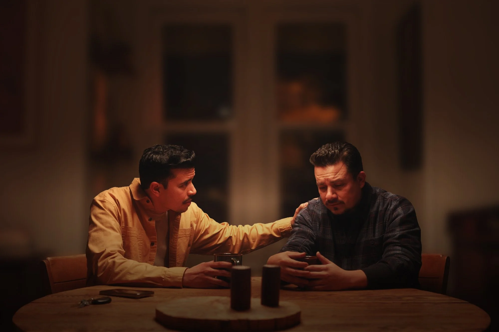 Two men sitting at a wooden table in a dimly lit room, one is holding the other's shoulder, and they are engaged in conversation.
