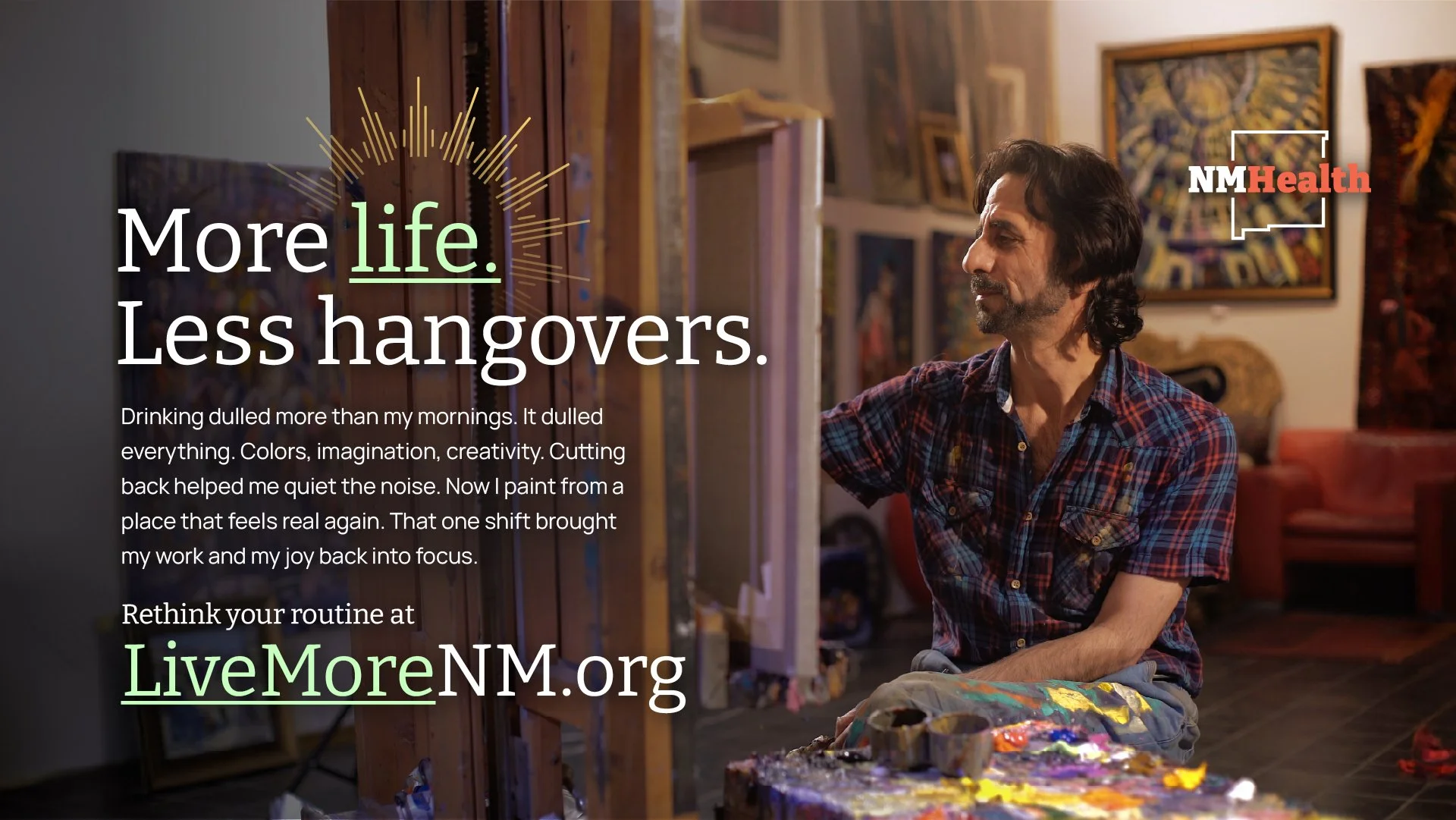 A man with dark hair and a beard, wearing a plaid shirt, is painting at an easel in an art studio. The background features colorful artwork and framed paintings on the wall, accompanied by the LiveMoreNM.org logo and text promoting healthier living.