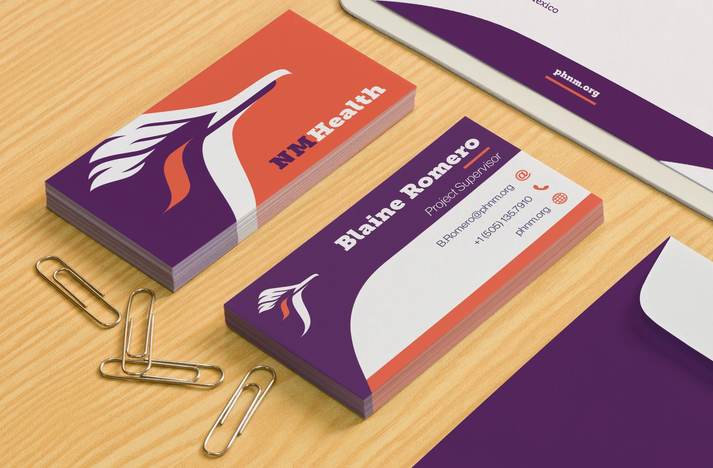Business cards on a wooden desk, branding for 'MHMHealth' and 'Blain Romero Project Supervisor,' with purple, orange, and white color scheme, paperclips, and a brochure partially visible.
