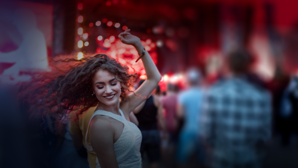 A woman dancing and smiling at a party or concert with people and colorful lights in the background.