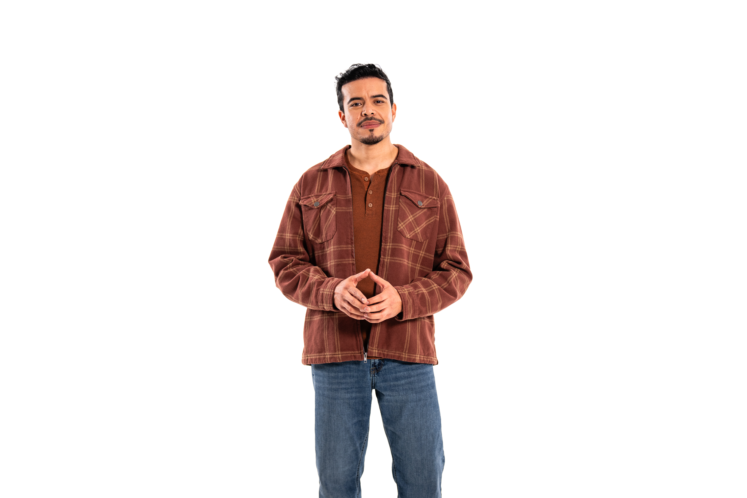 A man with dark hair, facial hair, wearing a brown plaid jacket over a rust-colored shirt and blue jeans, standing with his hands clasped in front of him against a plain background.