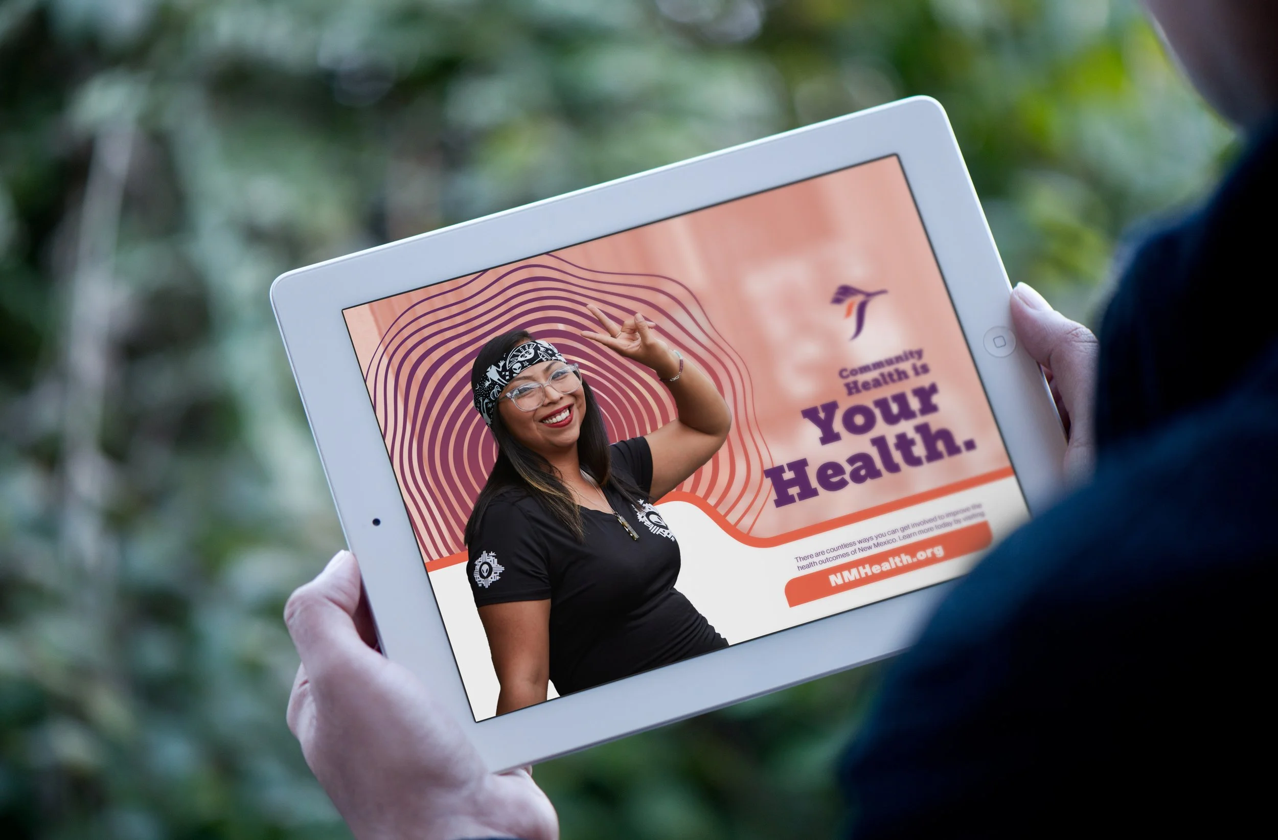 A person holding a white tablet displaying a colorful health awareness webpage featuring a smiling woman with glasses and a headband, with the text 'Community Health is Your Health.' The background is blurred with greenery.