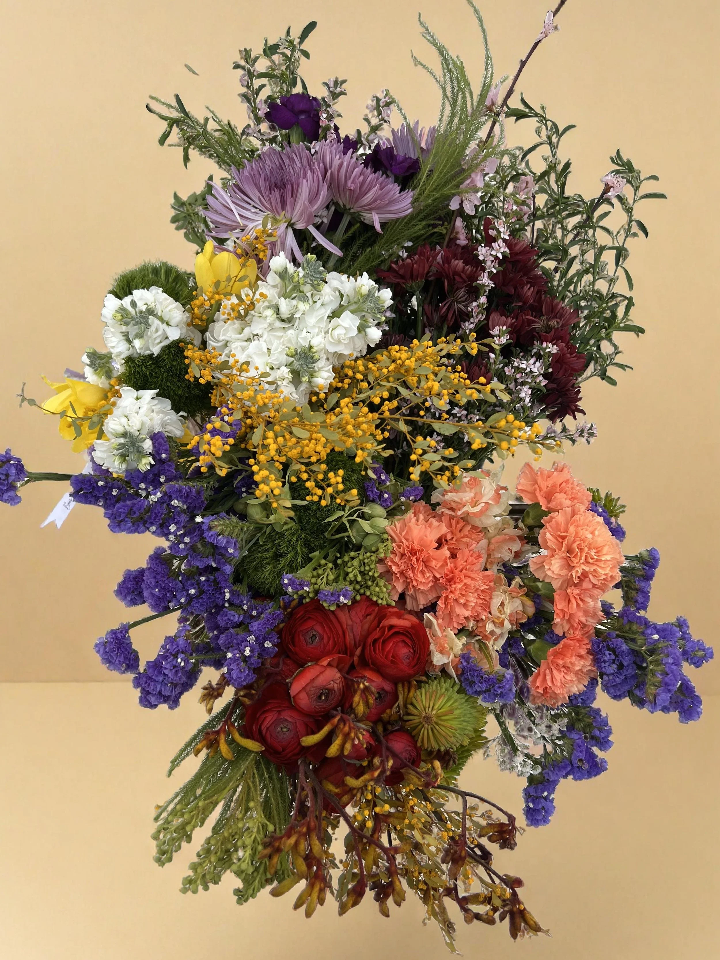 A colorful bouquet of various flowers including purple, white, yellow, red, and orange blooms against a plain beige background.