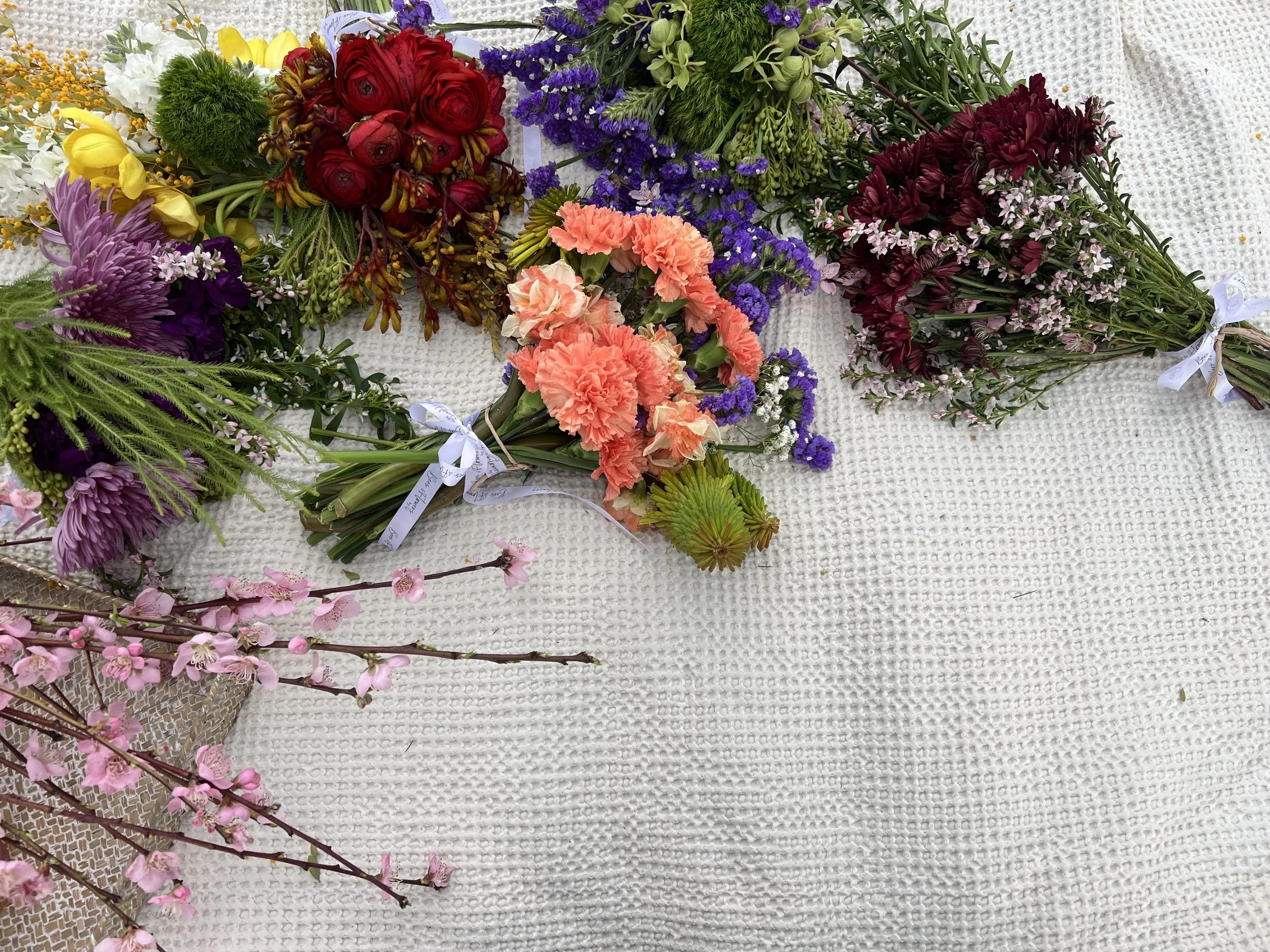 Various colorful flower bouquets, including red, purple, pink, yellow, and white flowers, arranged on a white textured surface, some with ribbons tied around the stems.