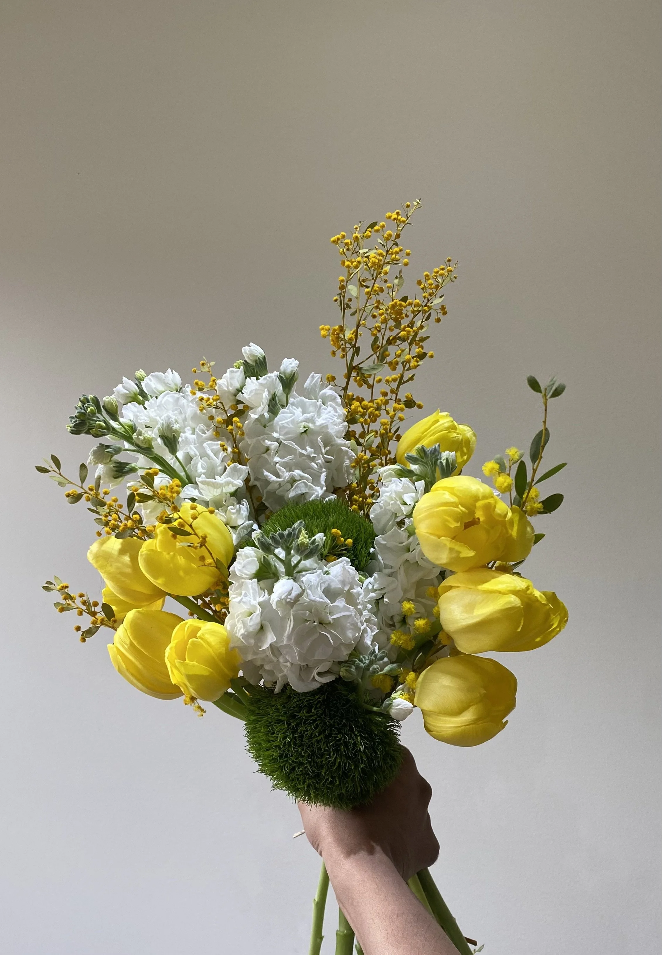 A hand holding a bouquet of yellow and white flowers, including tulips and other blooms, against a plain, light-colored background.