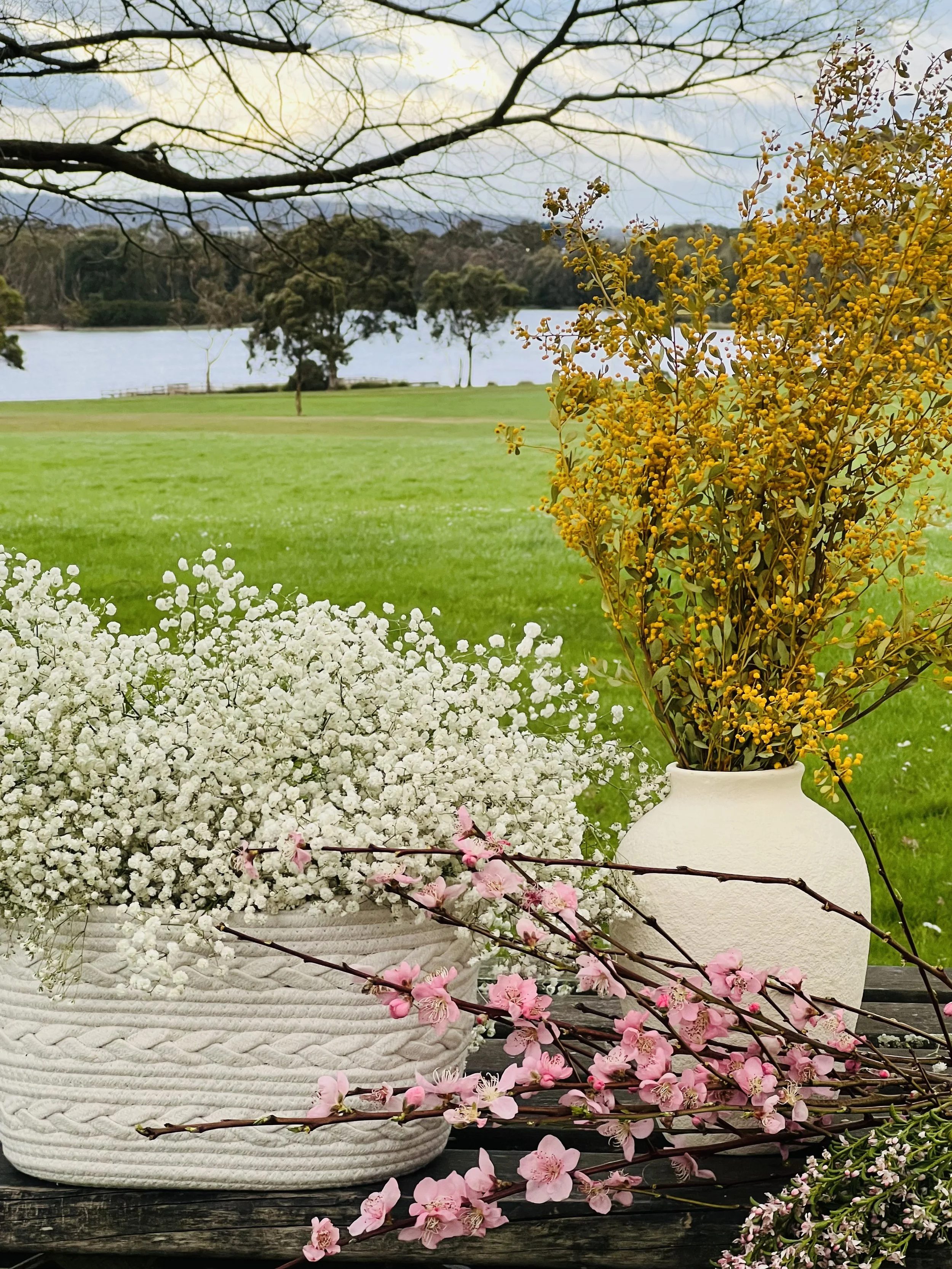 Vase with white baby's breath, vase with yellow flowers, and cherry blossom branch on wooden table outdoors with lake, trees, and cloudy sky in background.