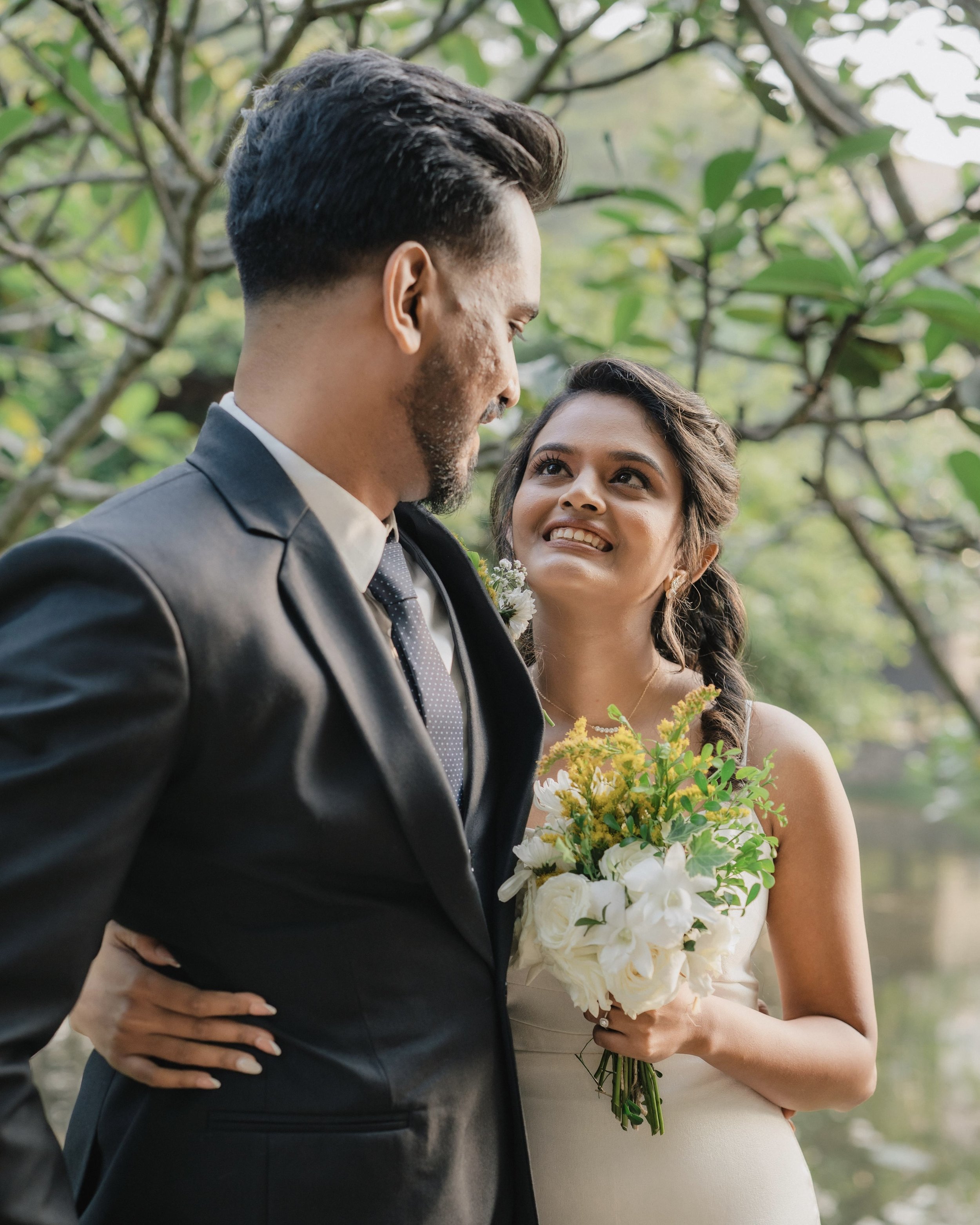 A bride and groom standing outdoors surrounded by greenery, gazing at each other. The bride is holding a bouquet of white flowers and greenery, wearing a white dress and earrings. The groom is in a dark suit with a tie. The scene is romantic and natural.