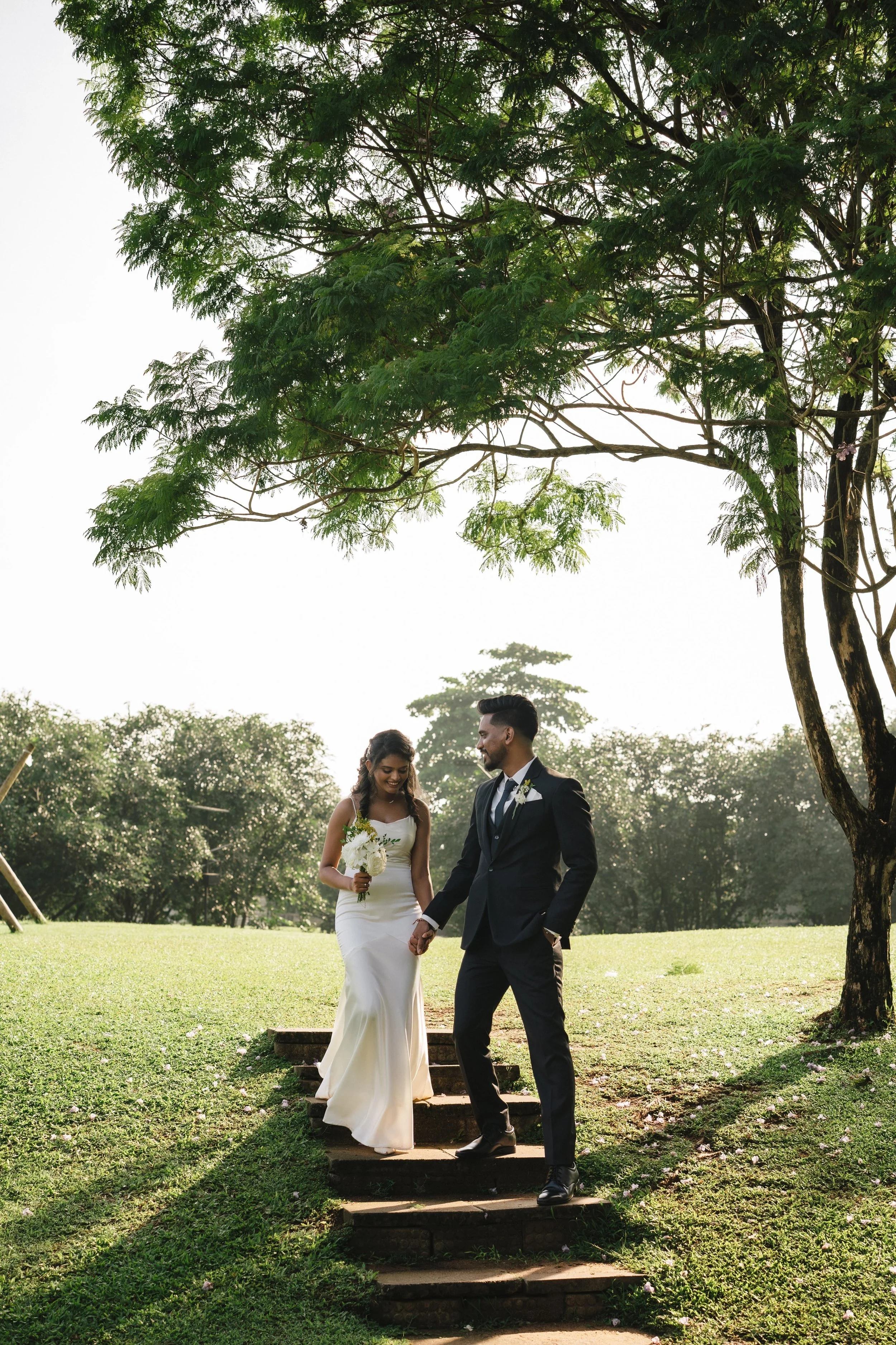 A bride and groom holding hands and walking down steps outdoors in a park, with trees and green grass, on a sunny day.