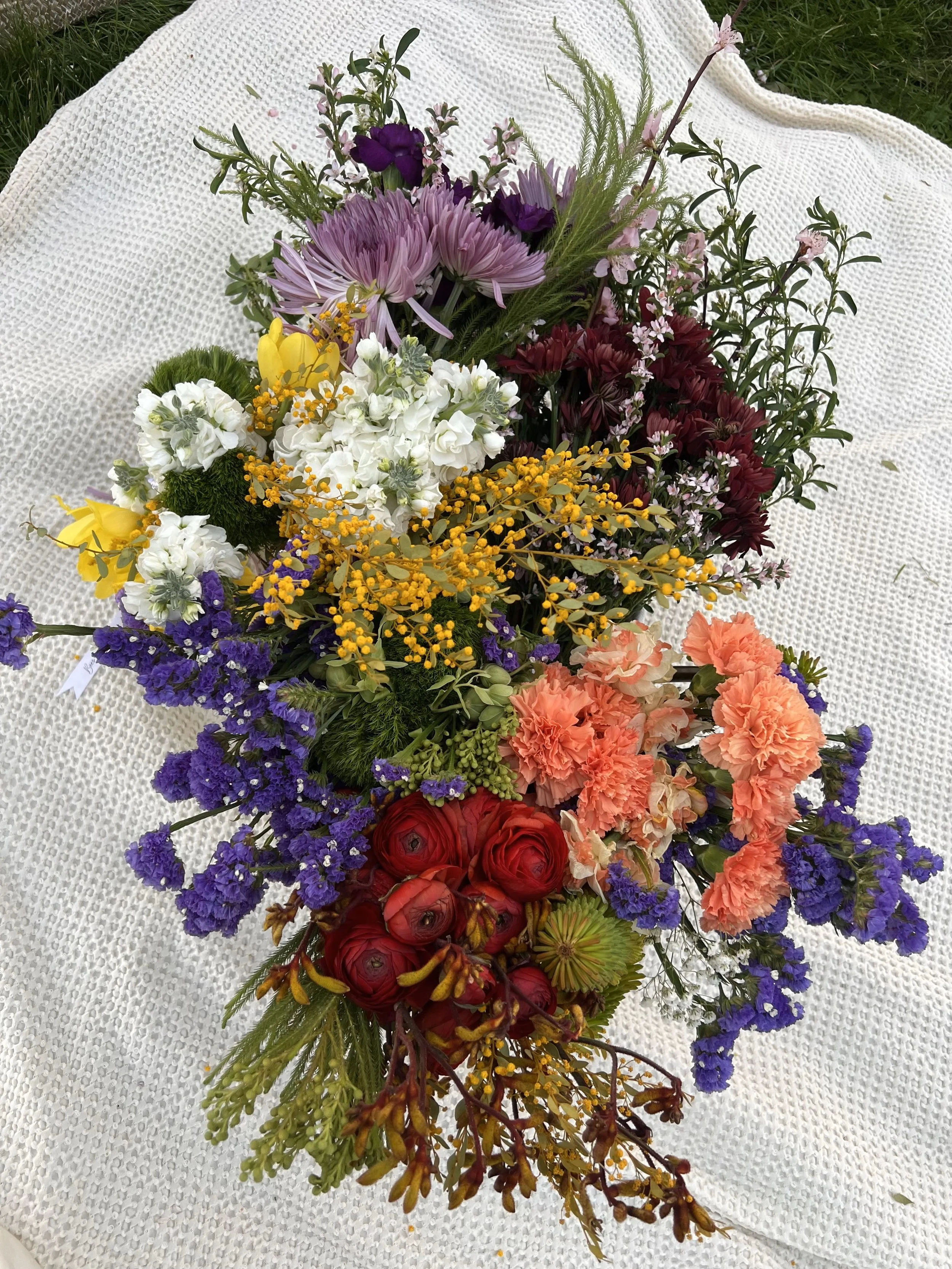 A colorful bouquet of various flowers including purple, yellow, white, red, peach, and violet blooms, resting on a textured white fabric surface with green grass visible in the background.
