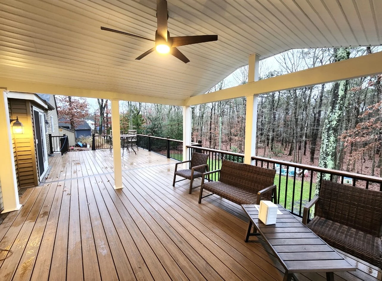 Covered deck with wicker furniture, ceiling fan, and view of wooded area.