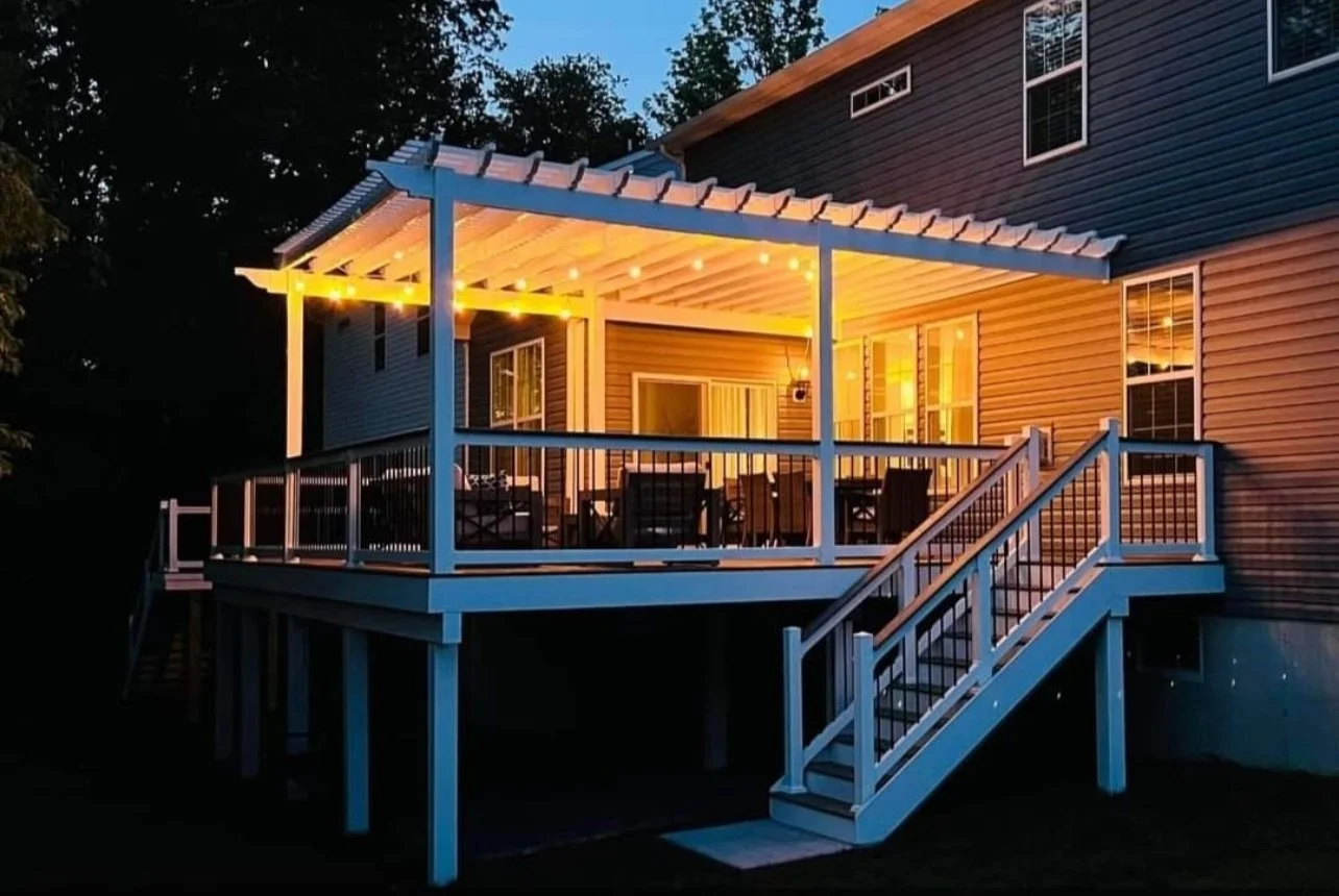 Outdoor deck with pergola and string lights attached to a house, featuring a railing and stairs leading down to the yard.