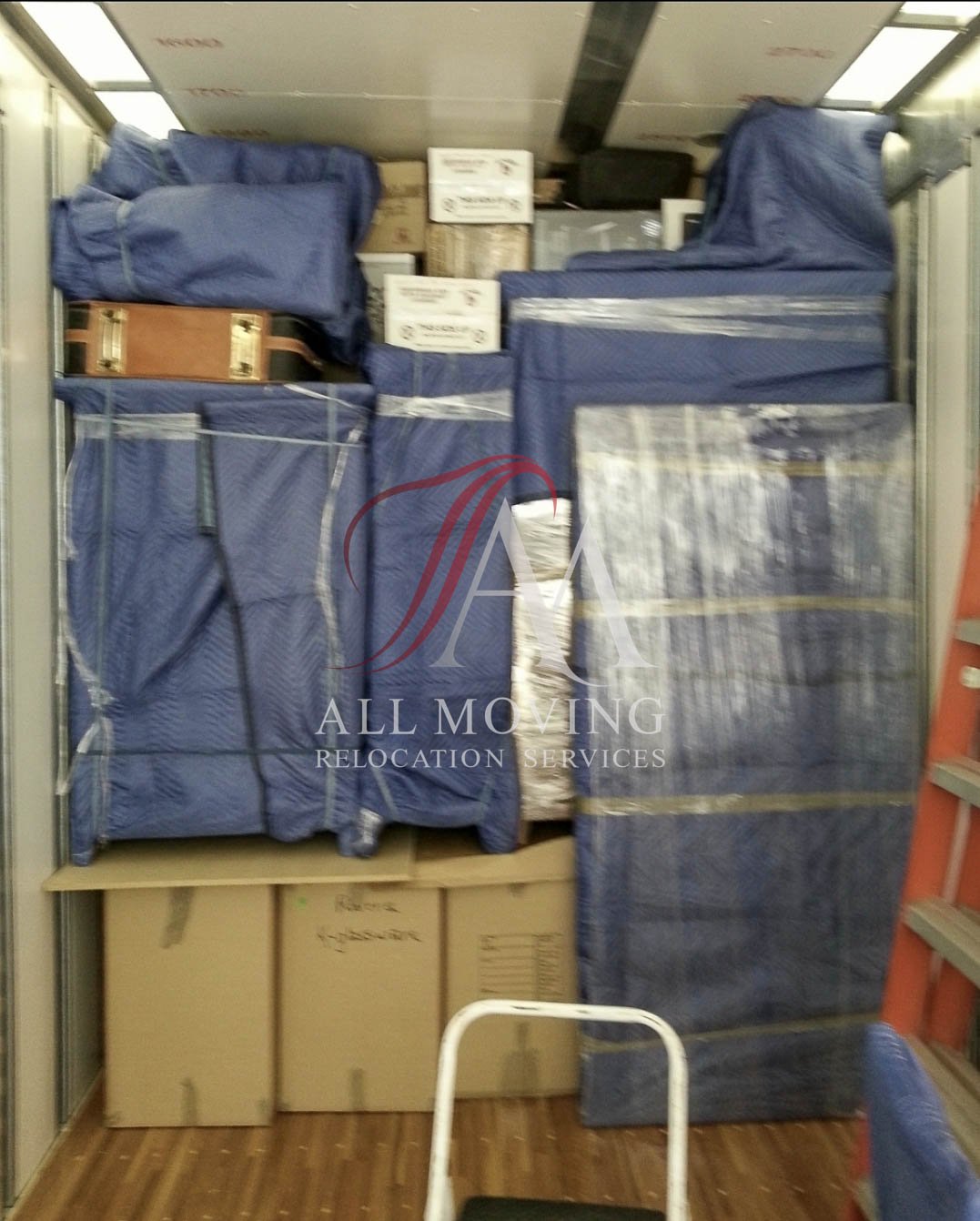 Packed moving truck with boxes and furniture covered in blue moving blankets, featuring "All Moving Relocation Services" branding.