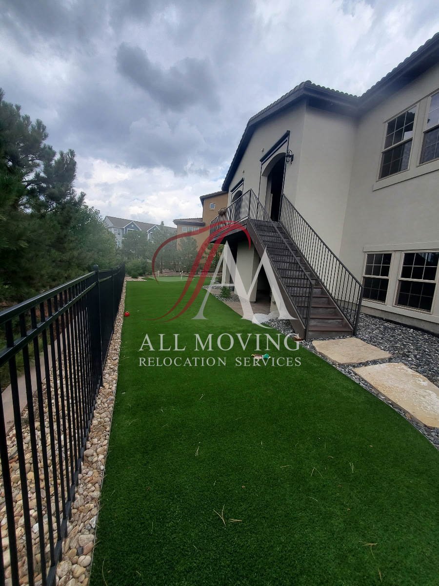 Outdoor view of a house with a staircase leading to a door, next to a fenced yard with artificial grass, under a cloudy sky. Branding for All Moving Relocation Services is overlaid on the image.