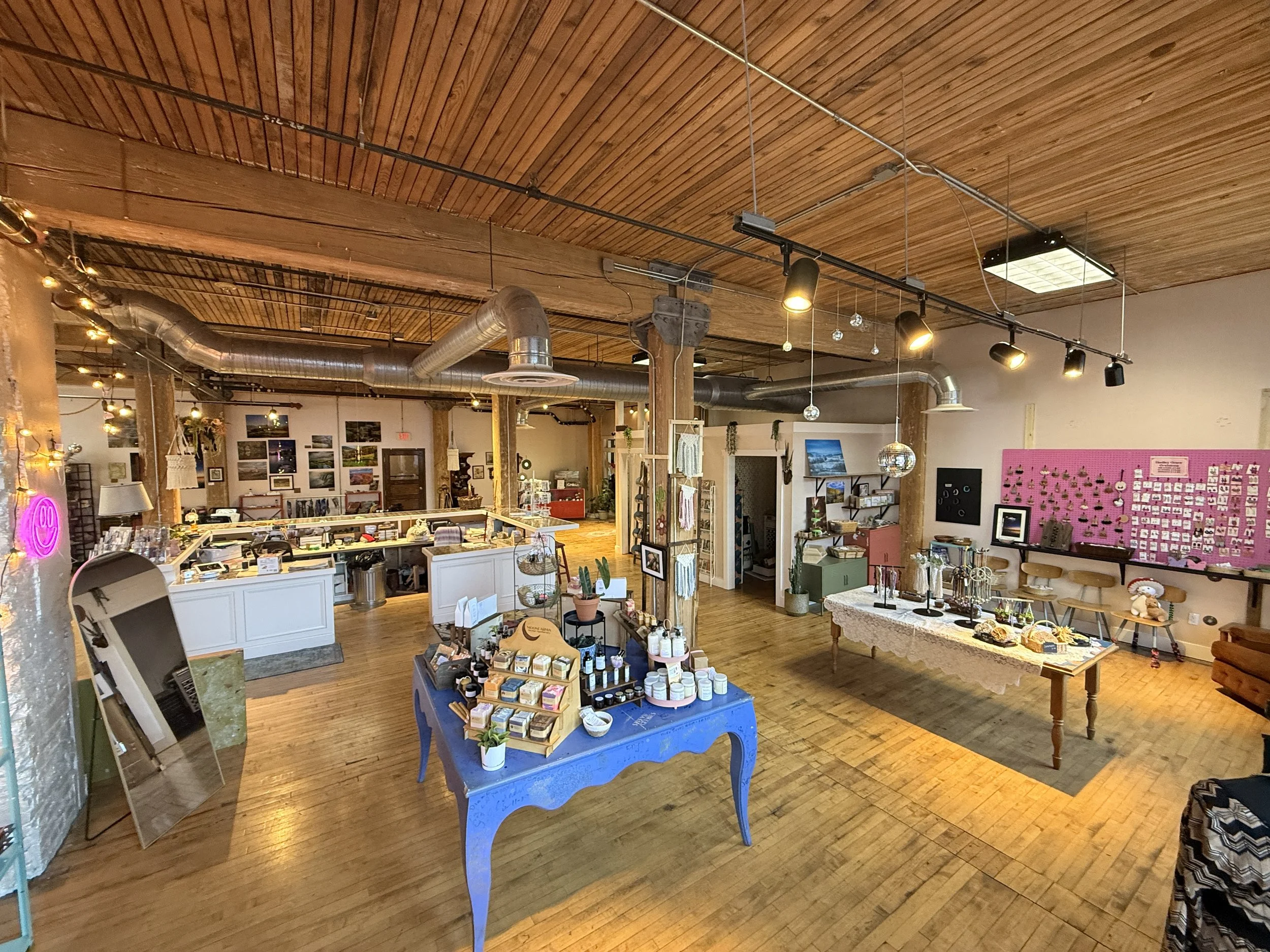 Interior of a boutique shop with wooden floors and ceiling, displaying jewelry, crafts, and home decor items on tables, shelves, and a pink display wall.