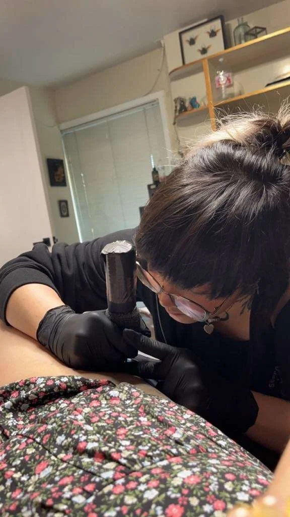 A tattoo artist in black gloves tattoos a clientβs arm in a cozy studio filled with framed art and shelves.