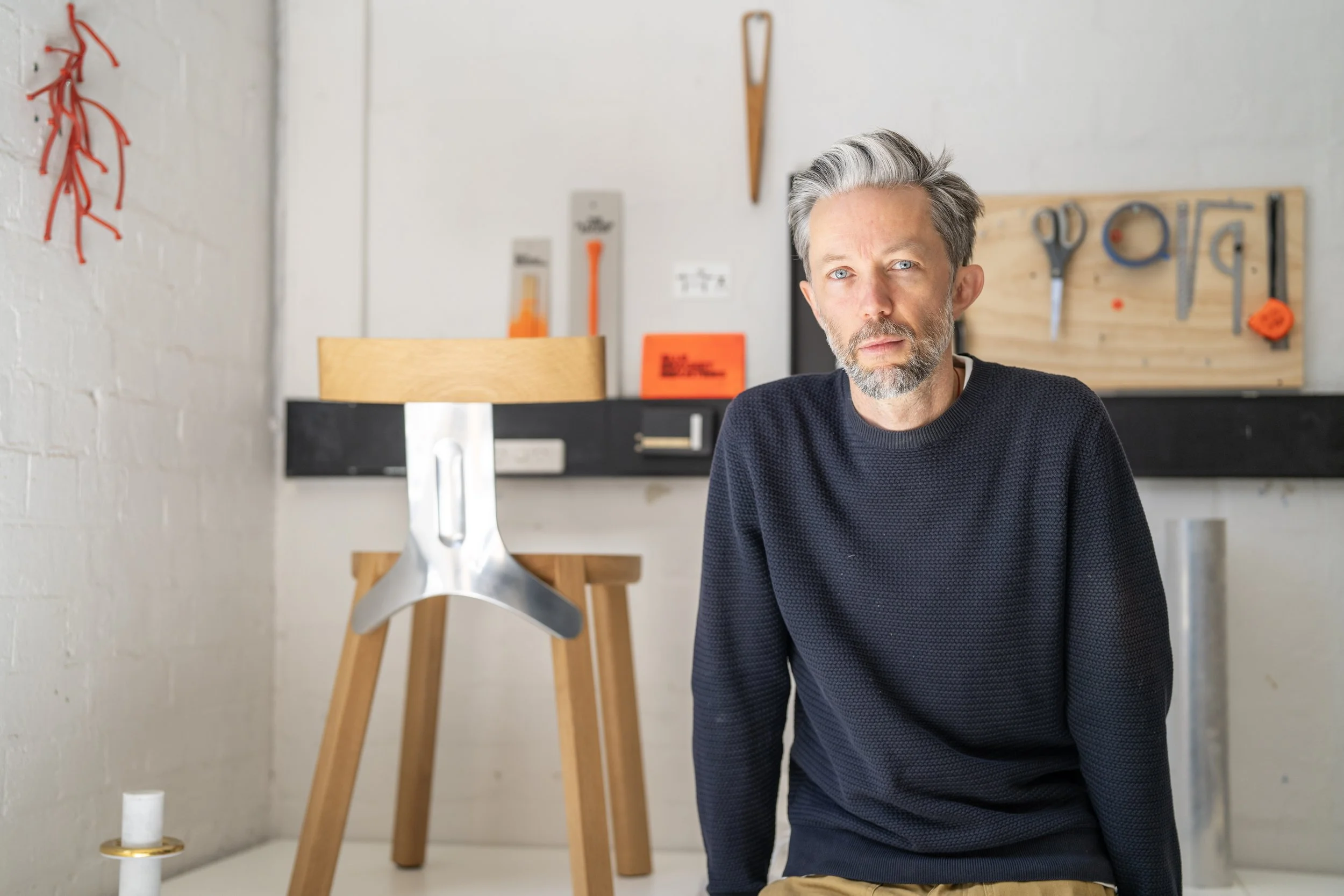 A man with gray hair and a beard, wearing a dark sweater, looks at the camera in a workshop with tools on a pegboard and a wooden chair.