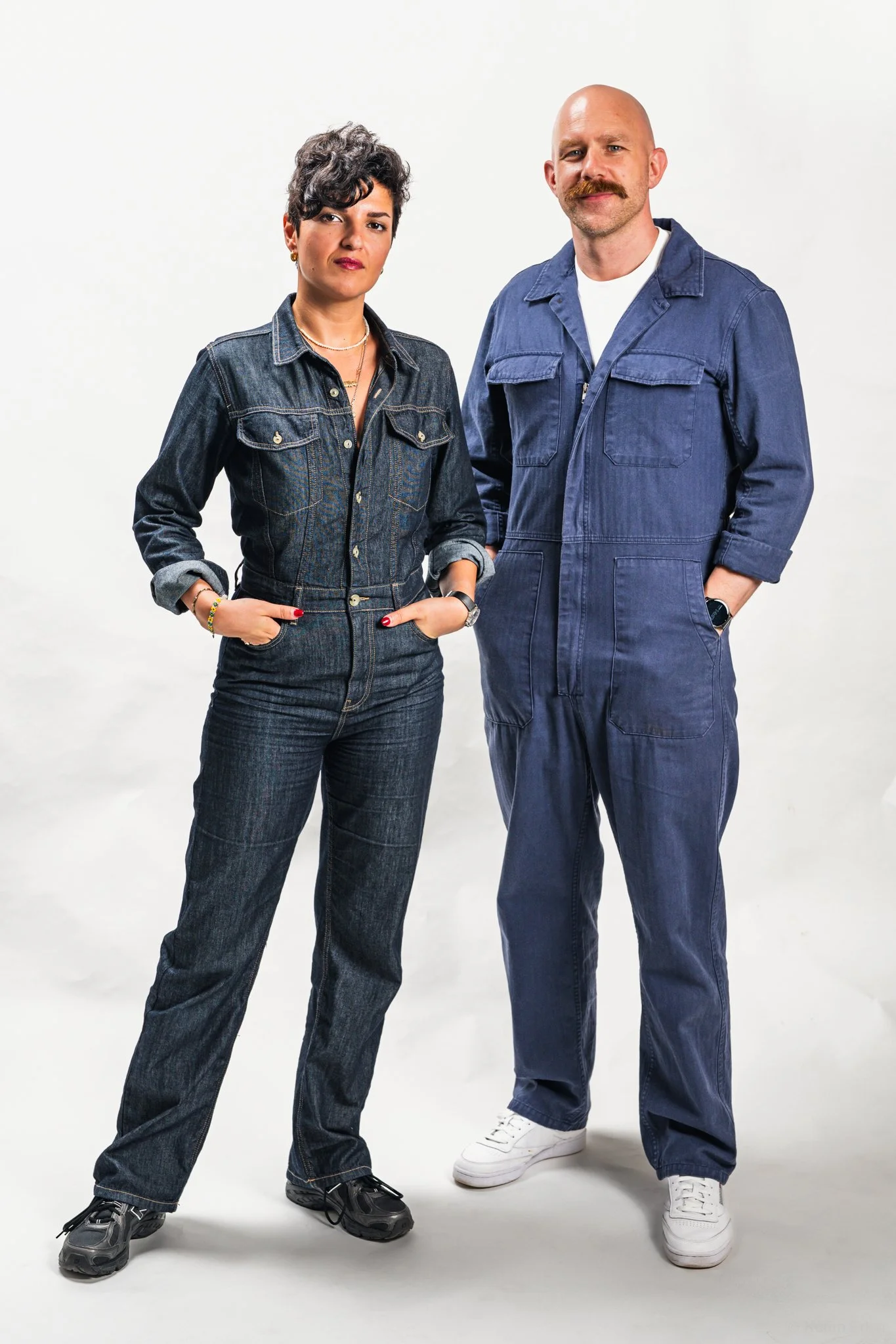 A woman and man standing against a plain white background, both wearing denim jumpsuits and looking at the camera.