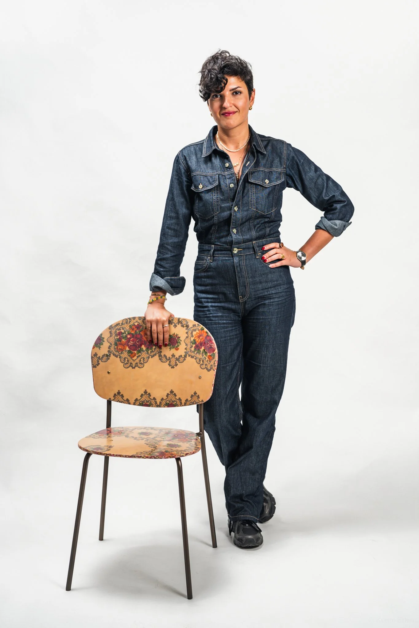 Woman in denim outfit standing next to an ornate chair with floral upholstery.