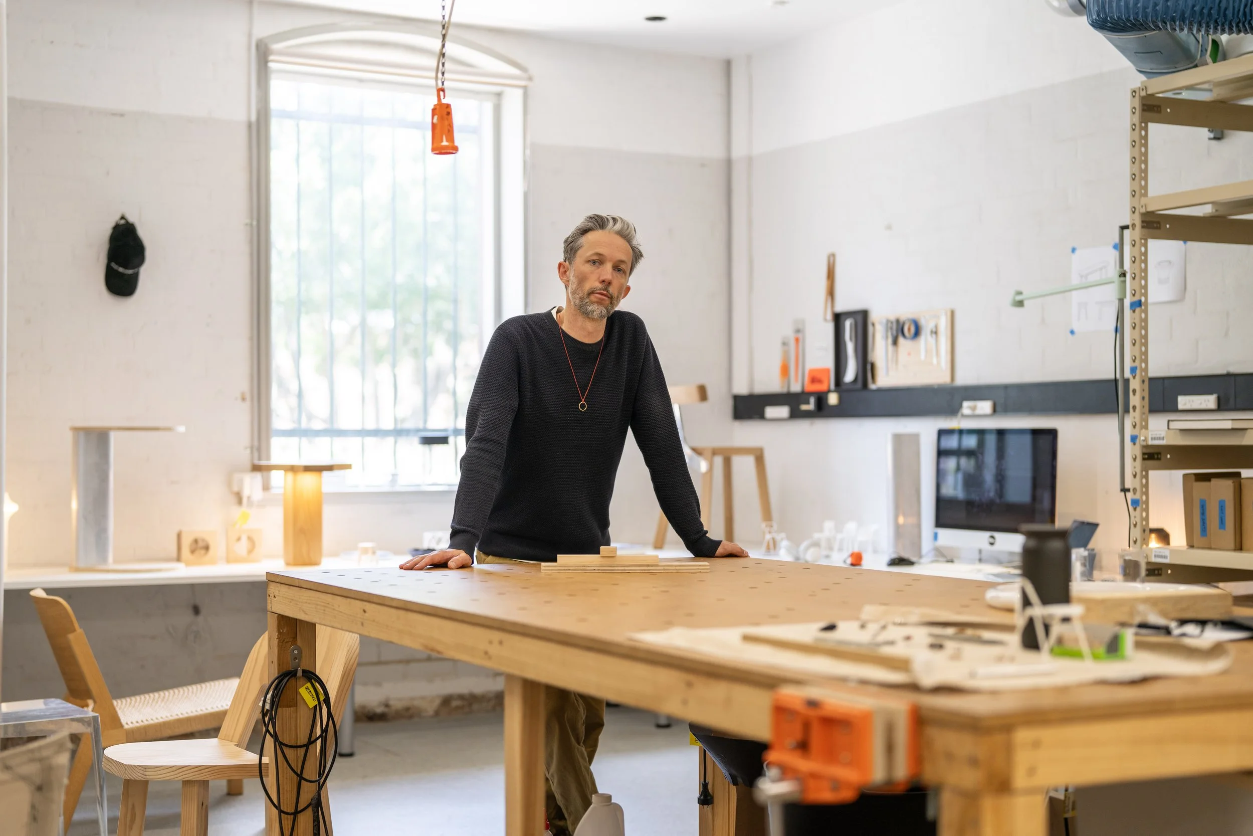 A man with gray hair and a beard standing in a woodworking workshop, leaning on a large wooden worktable with woodworking tools and equipment around him.