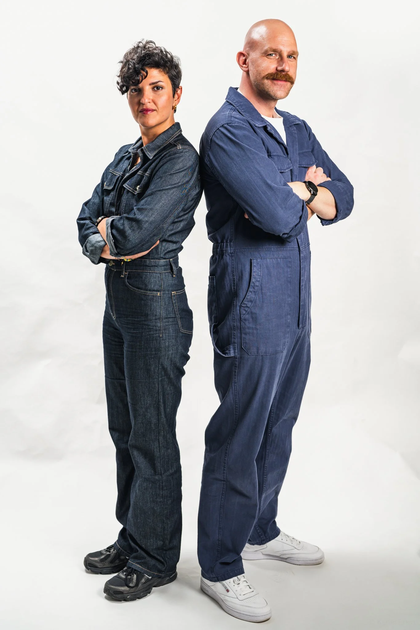 A man and woman standing next to each other with arms crossed, both wearing blue denim outfits, posing in front of a plain white background.
