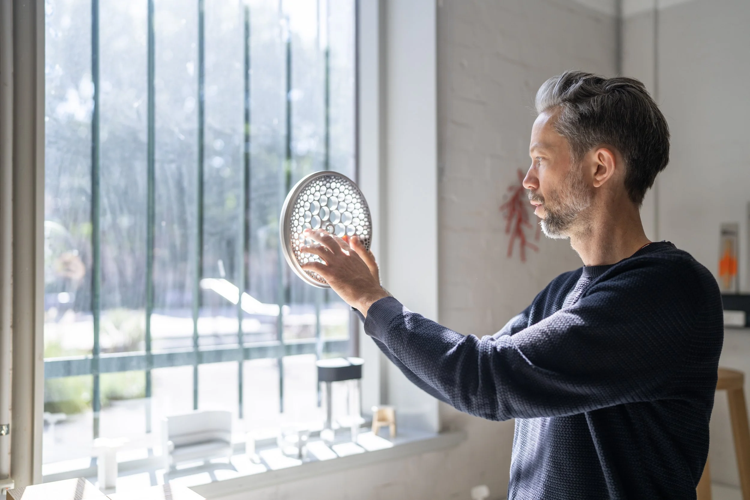 A man with gray hair and beard holding a circular object with holes near a large window, with sunlight streaming in.