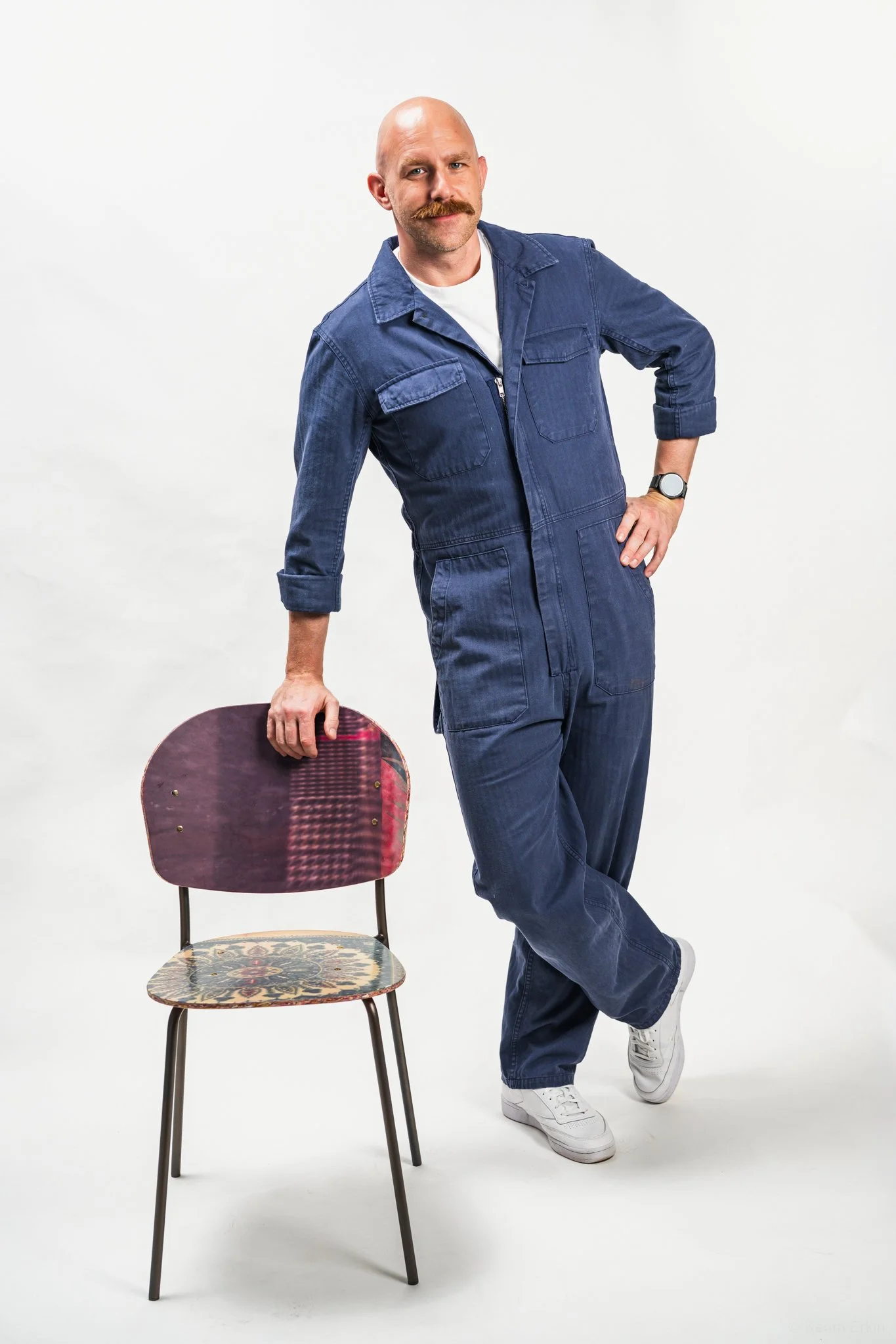 A man with a mustache wearing a blue jumpsuit, standing next to a vintage-style chair with a red and purple seat and a patterned cushion, in a studio setting with a plain white background.