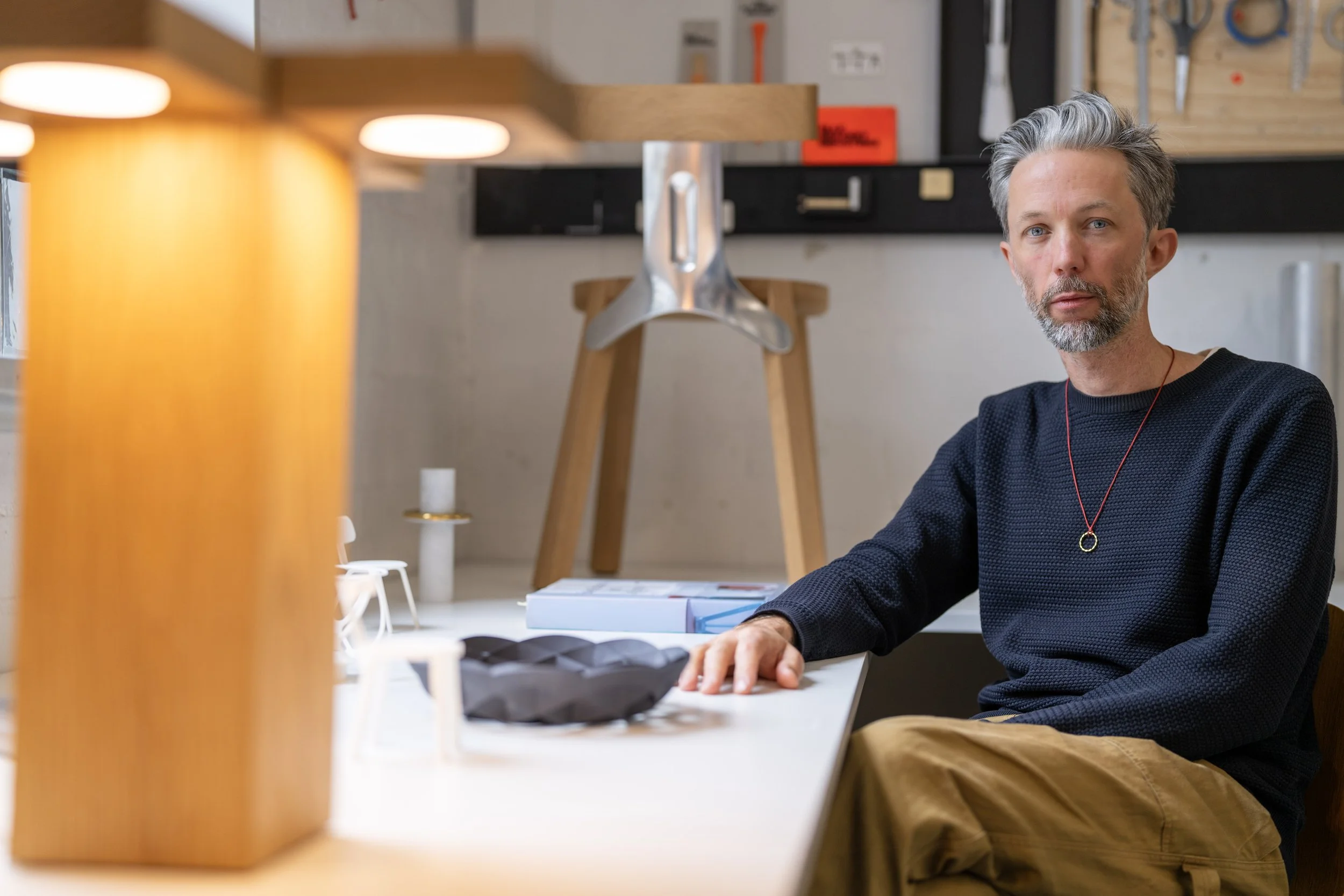 A man sitting at a desk in a workshop or studio, with tools on the wall behind him and a wooden table in front, partially obscured by a yellow object in the foreground.