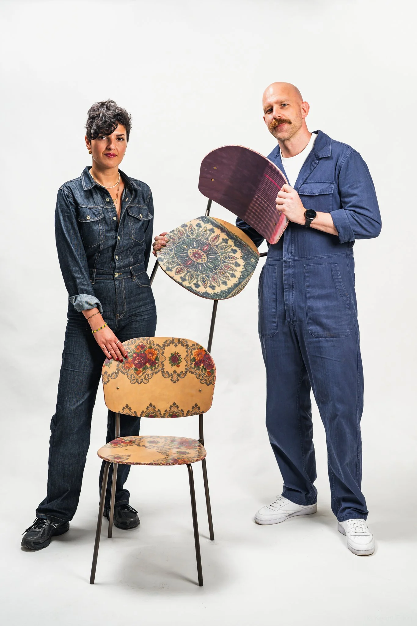 A woman and a man standing next to a chair with three different decorative chair backs balanced on its backrest. The woman has short, curly hair, wears a denim jumpsuit, and the man is bald with a mustache, dressed in a denim jumpsuit, holding a deco