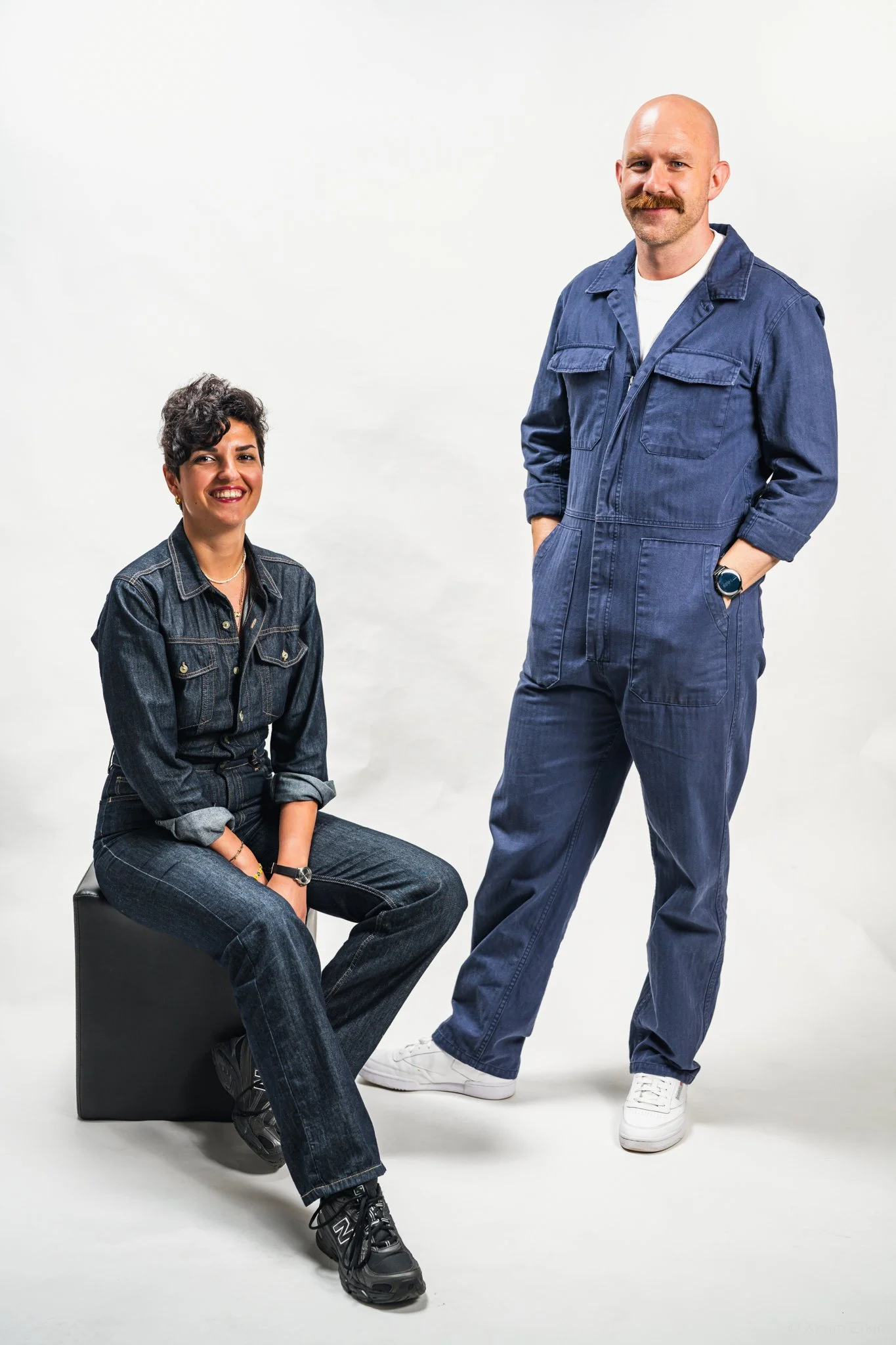 A woman sitting on a black box and a man standing against a plain white background. Both are wearing denim clothing and smiling.