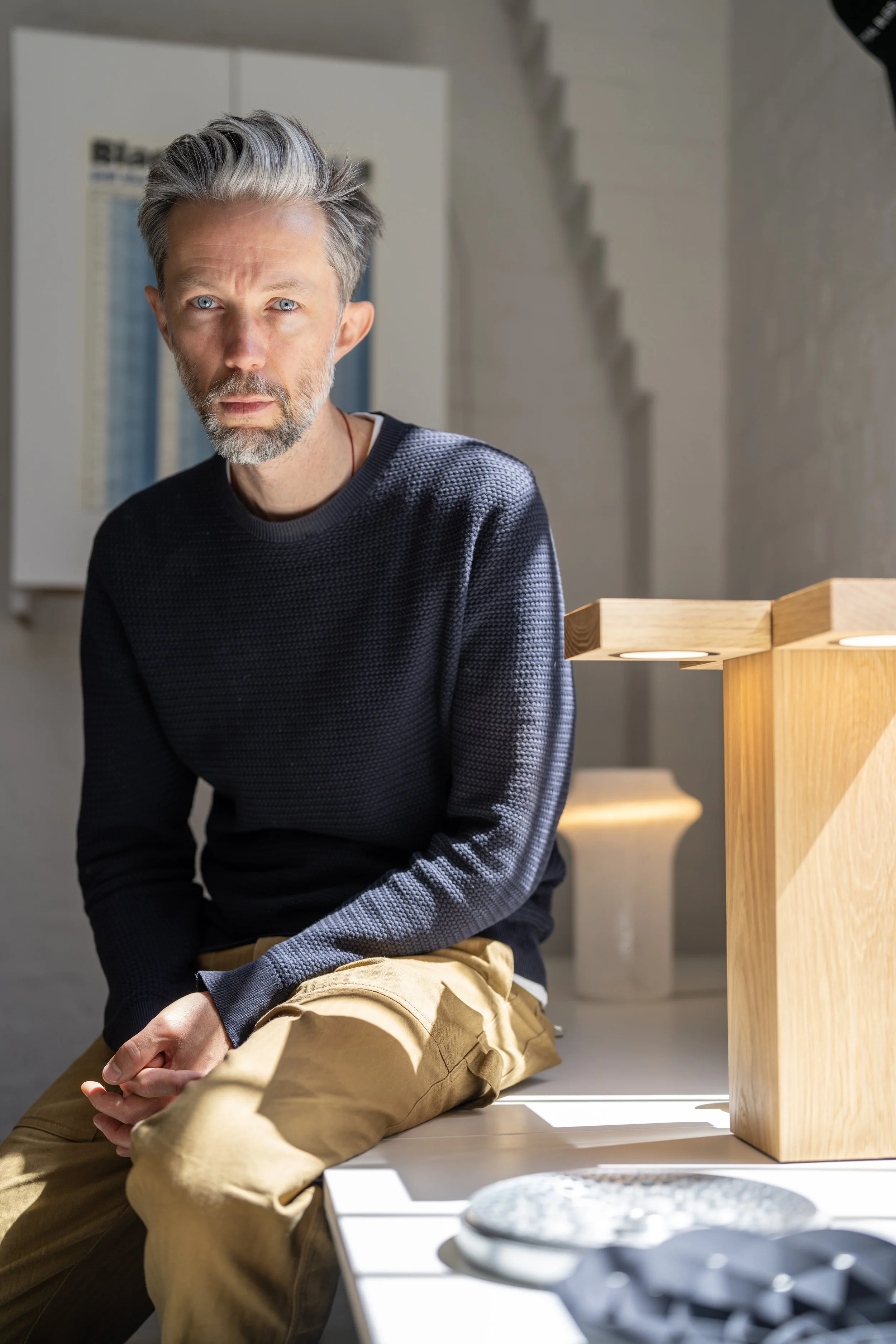 A middle-aged man with gray hair and a beard sitting on a white surface, wearing a black sweater and tan pants, in a bright room with sunlight streaming in.
