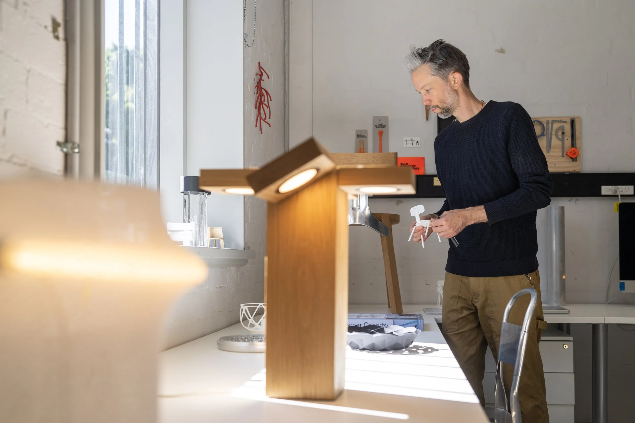 Man with gray hair and beard assembling furniture in a modern, well-lit workshop or studio with tools and decorations on the wall.