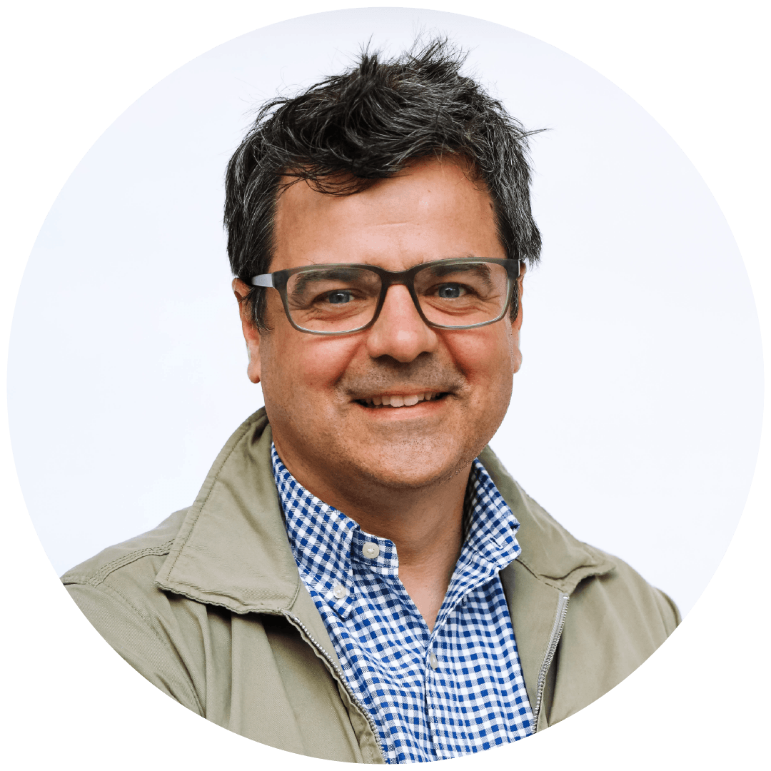 A man with dark, curly hair, wearing glasses, a beige jacket, and a blue checkered shirt, smiling against a white background.