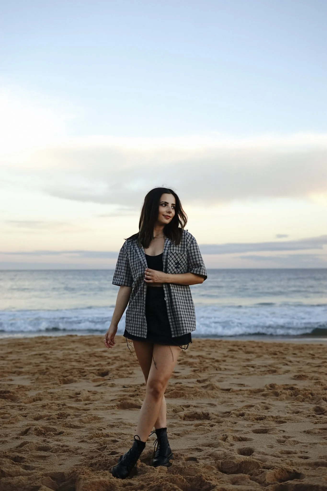 A young woman with dark hair wearing a plaid shirt, black top, and black shorts stands on a sandy beach during sunset, with the ocean and sky in the background. Copacabana Beach Central coast nsw
