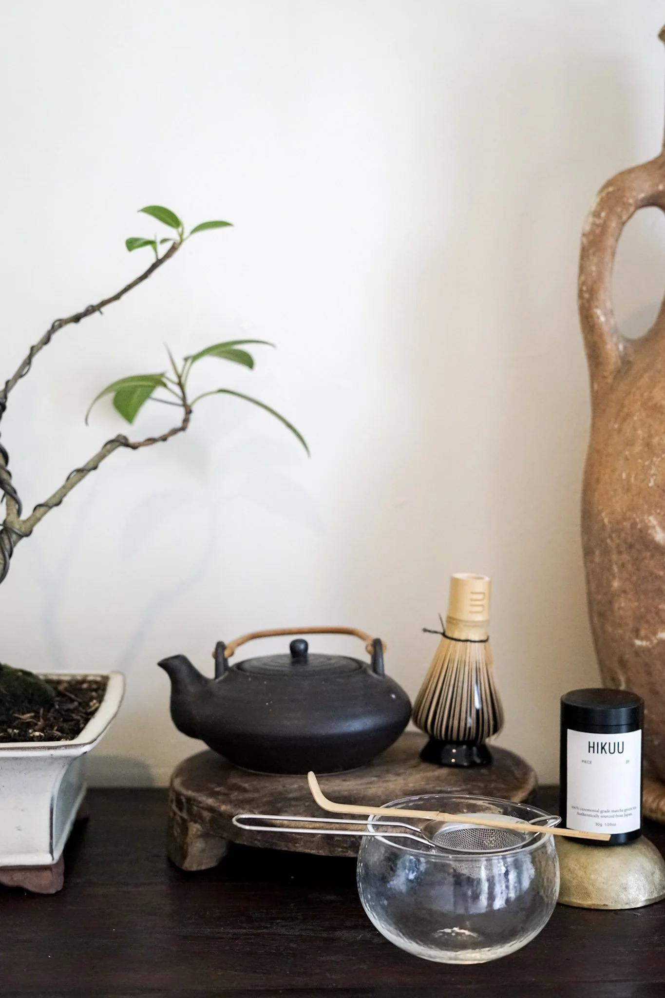 A collection of decorative items on a dark wooden surface, including a potted plant, a black teapot on a wooden stand, a clear glass bowl, a small bamboo brush, a black container labeled 'HIKUU', and a wooden stand with a black book. There is a white