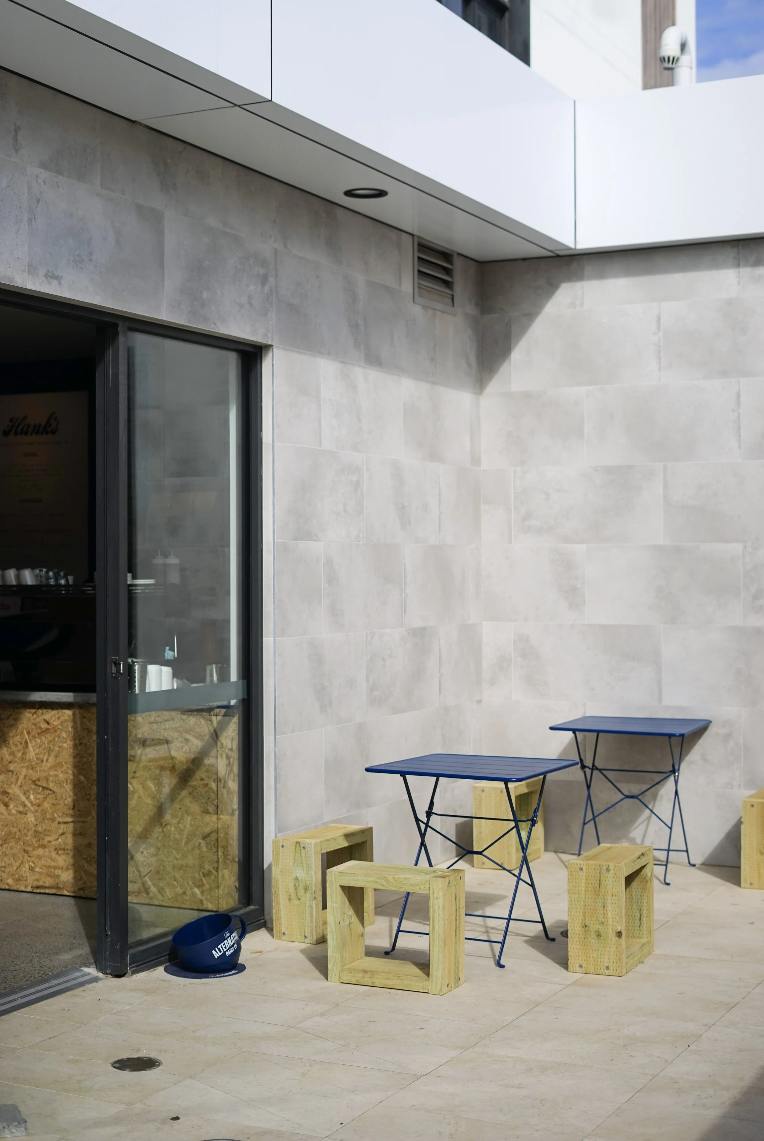 Outdoor seating area with two blue tables and wooden chairs, beige tiled floor, modern building wall with a window and ventilation grate, blue helmet on the ground, and a black and blue sign. Ettalong beach nsw