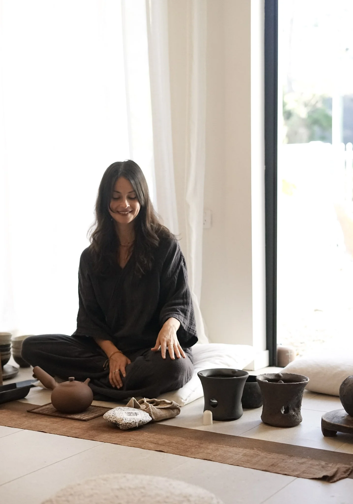 A woman with long dark hair, wearing a black robe, is sitting cross-legged on a tatami mat, involved in a Japanese tea ceremony with traditional tea utensils on a woven mat, indoors near a large window with sheer curtains.