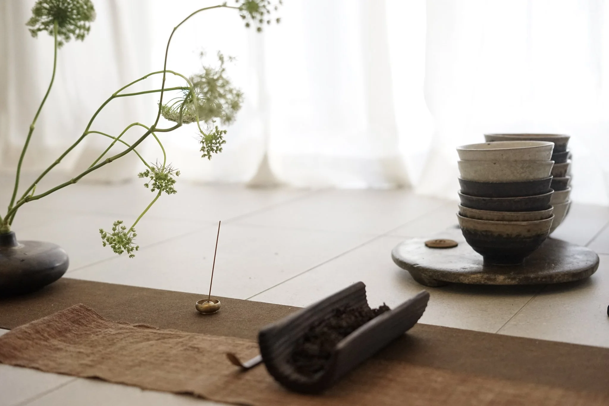A collection of ceramic bowls stacked on a slate tray, with a brown cloth underneath, and a small brass incense holder with a stick of incense in it, on a table with a brown tablecloth. In the background, there are vases with green plants and white c