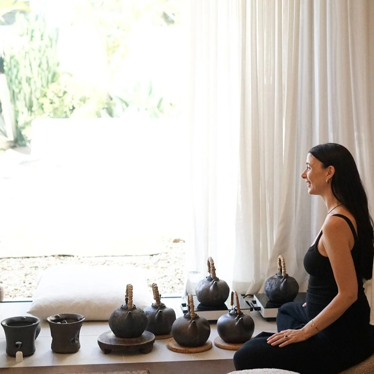 A woman sitting cross-legged near a table with several black pottery teapots and cups, smiling as she looks at a large window with white curtains. Outside, garden plants are visible.