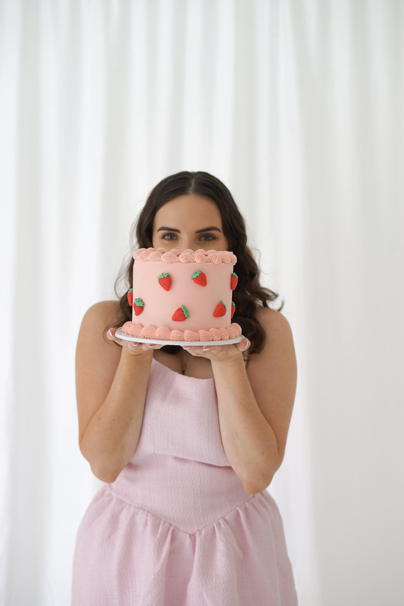 A woman in a pink dress holding a pink cake with strawberry decorations in front of her face, standing against a white background.