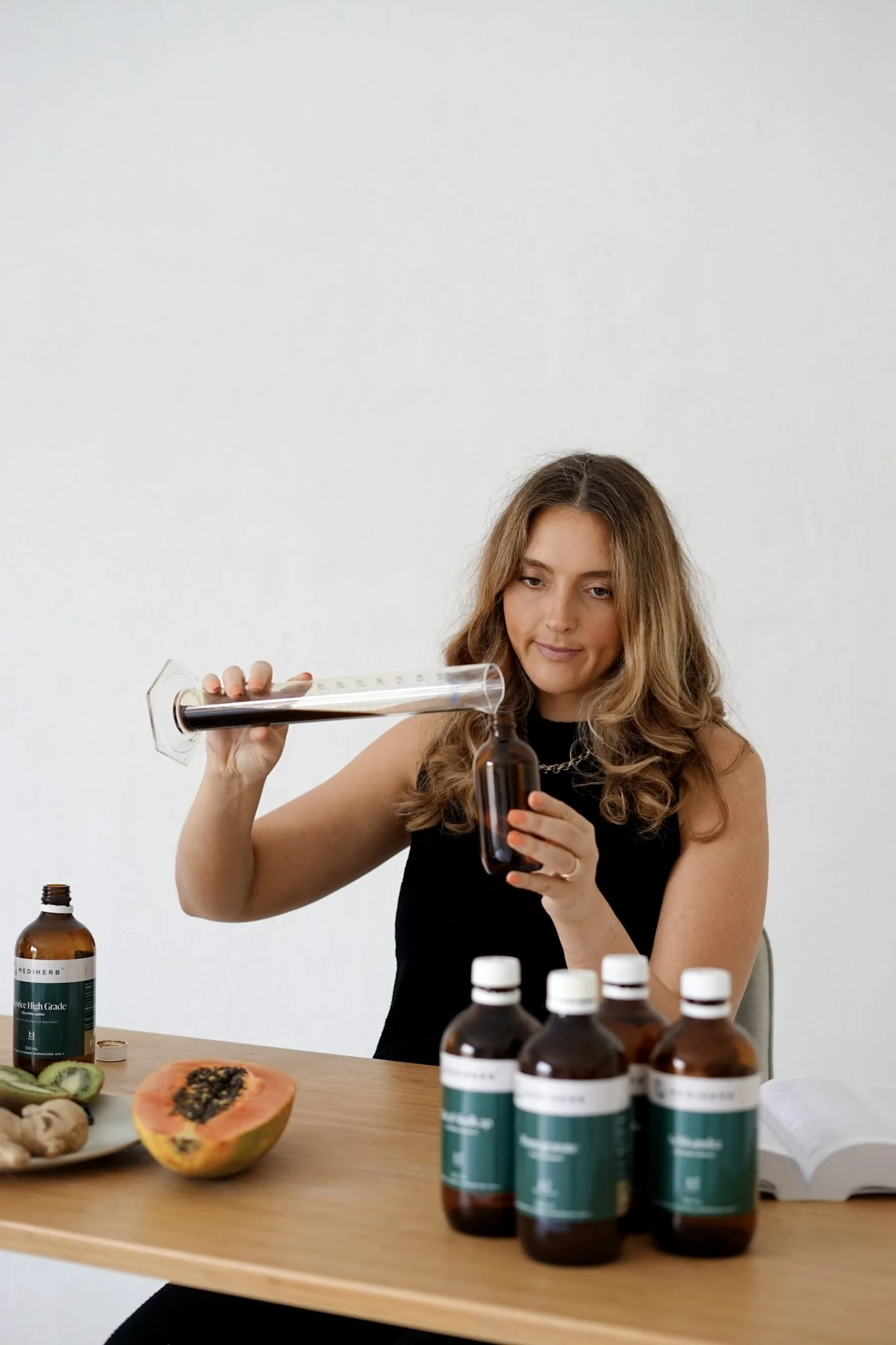 A woman with long wavy hair wearing a black top is sitting at a table, pouring a dark liquid from a test tube into a small brown bottle. The table has several bottles, a halved papaya with black seeds, sliced kiwi, ginger, and an open book. The backg