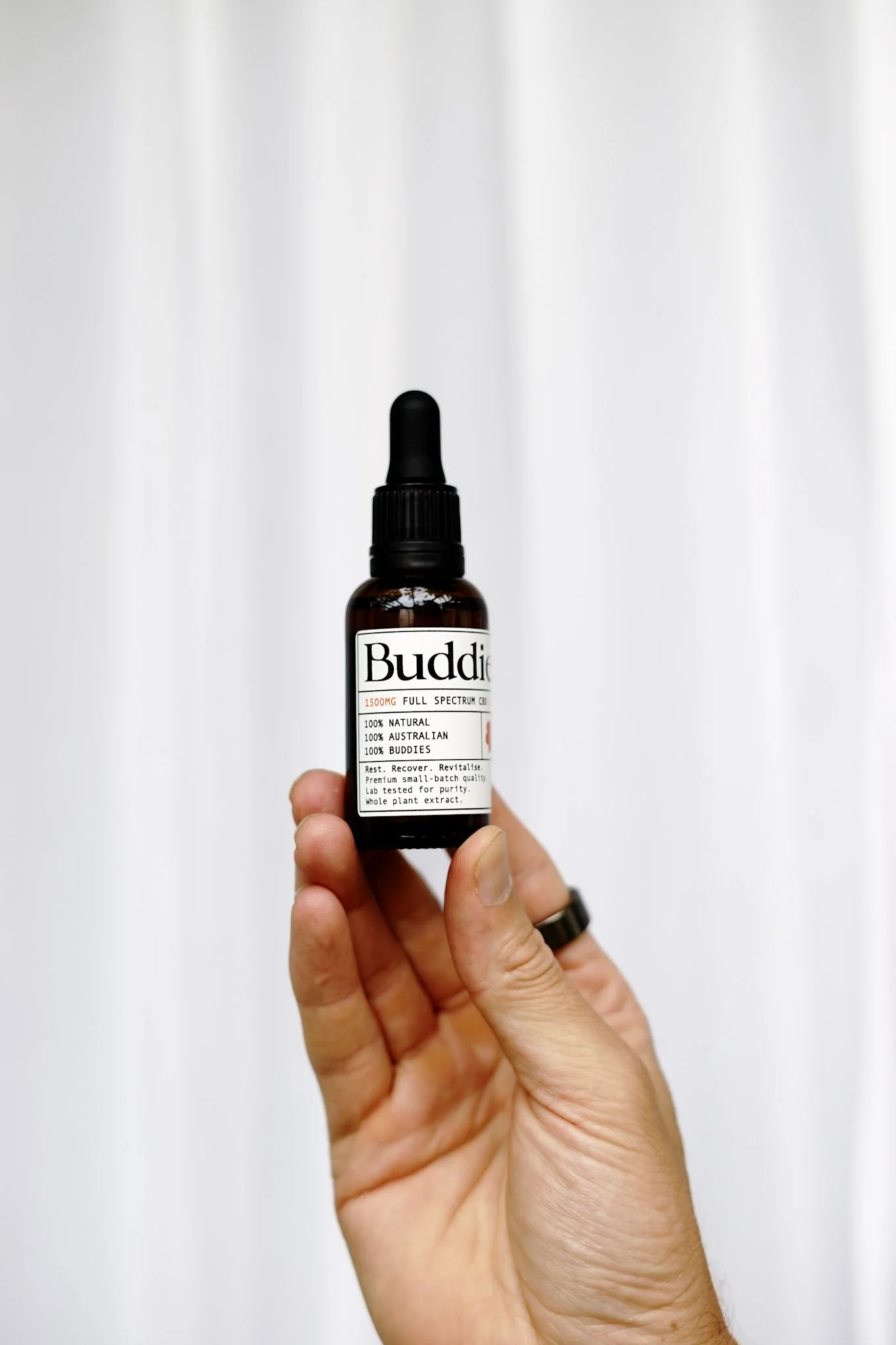 Hand holding a small brown bottle labeled 'Buddie' with a dropper cap, against a plain light background.