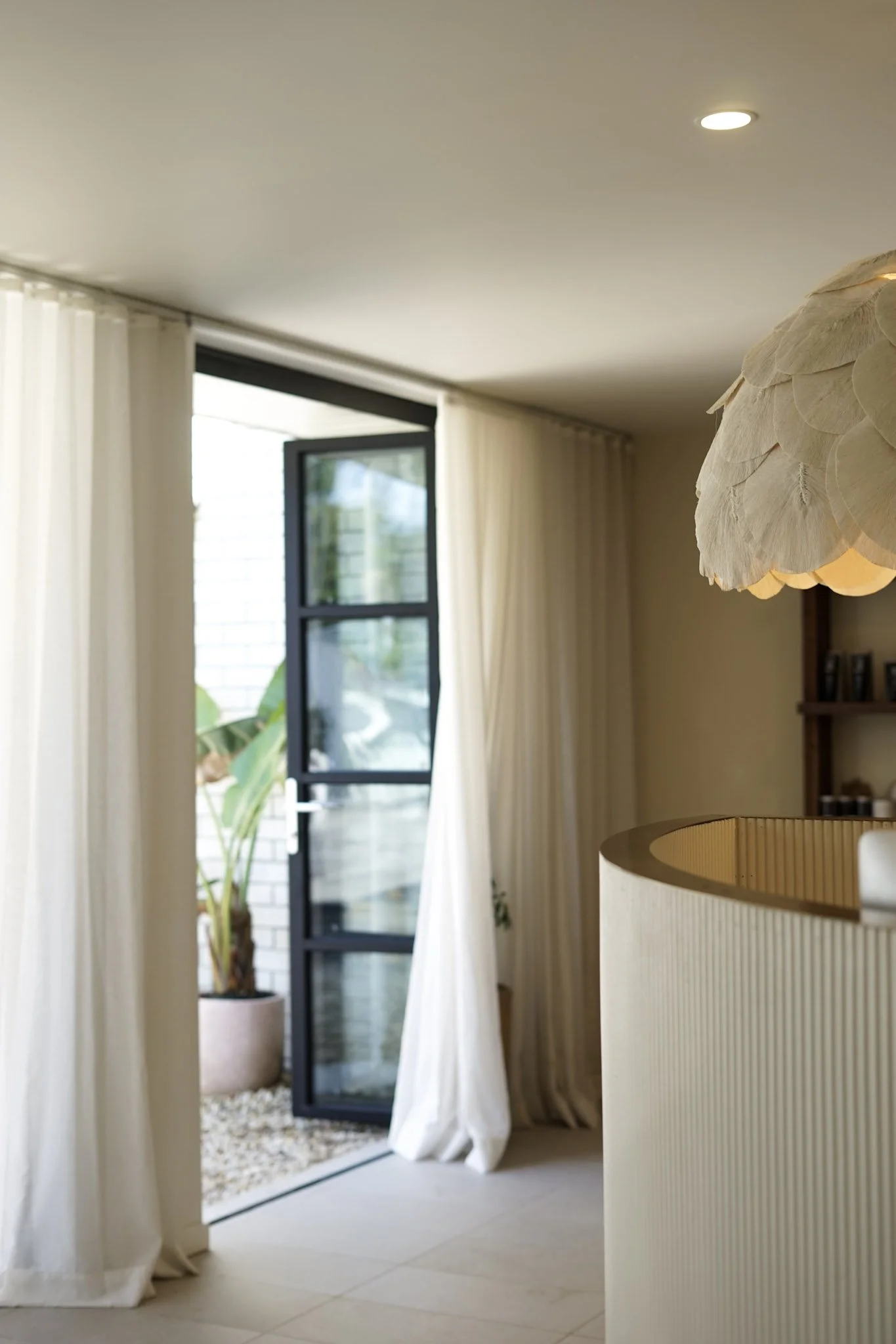Interior of a modern home with off-white curtains, a black-framed glass door open to an outside area with green plants, and a hanging light fixture with wood scales.