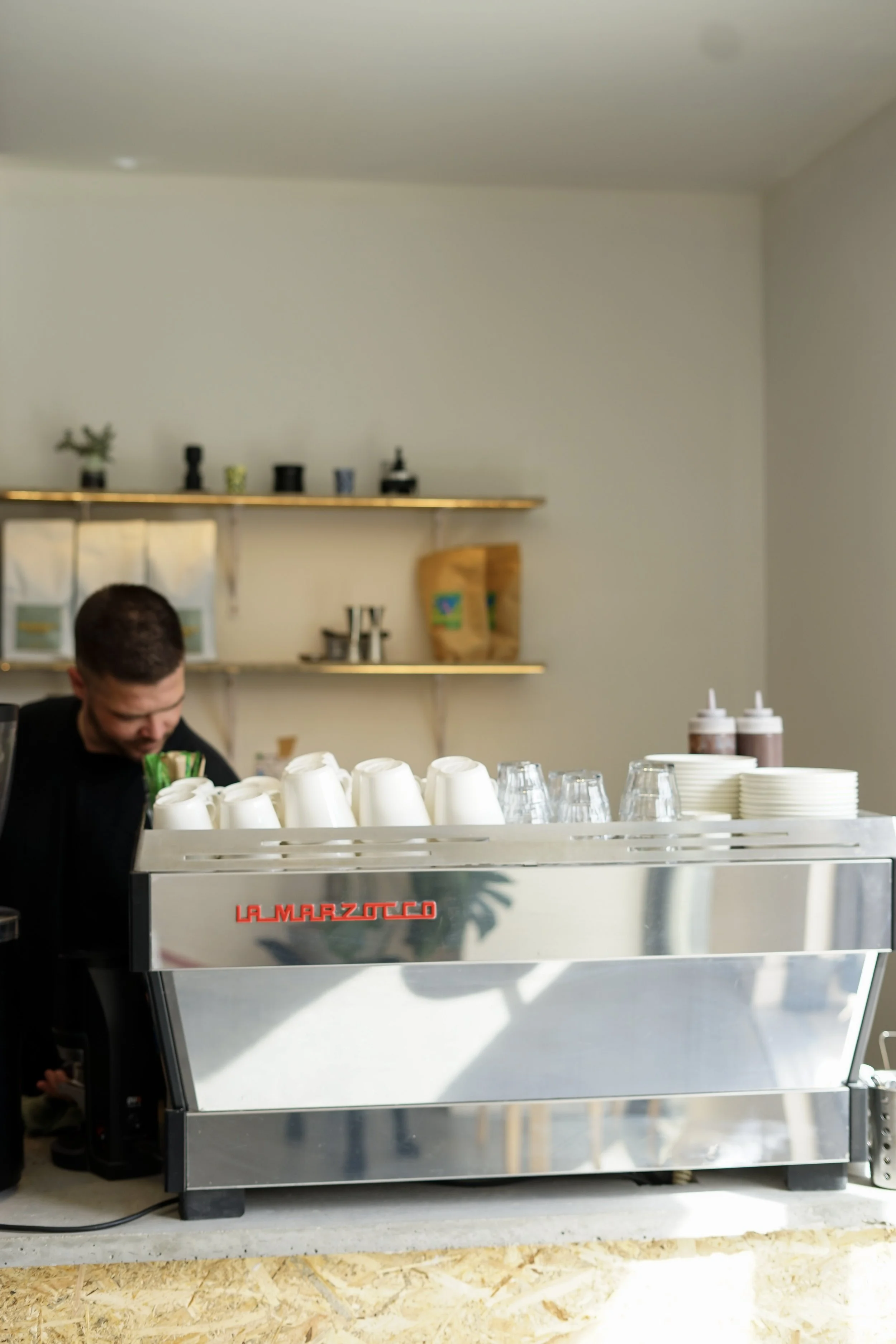 A person working behind a La Marzocco espresso machine in a cafe, with cups, glasses, and bottles on the counter and shelves with plants and supplies in the background.