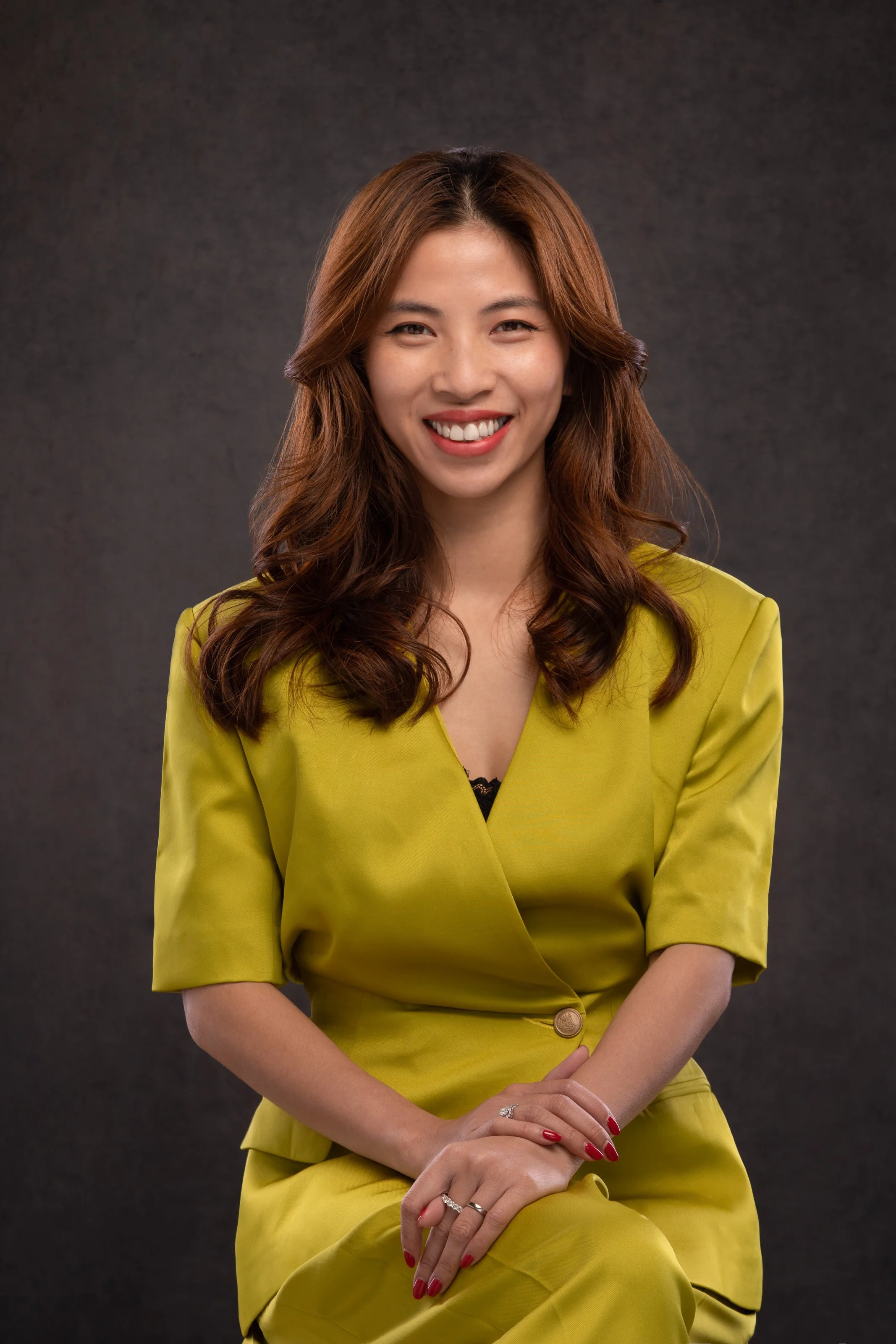 Smiling woman with wavy brown hair wearing a lime green blazer and matching skirt, sitting against a dark grey background.