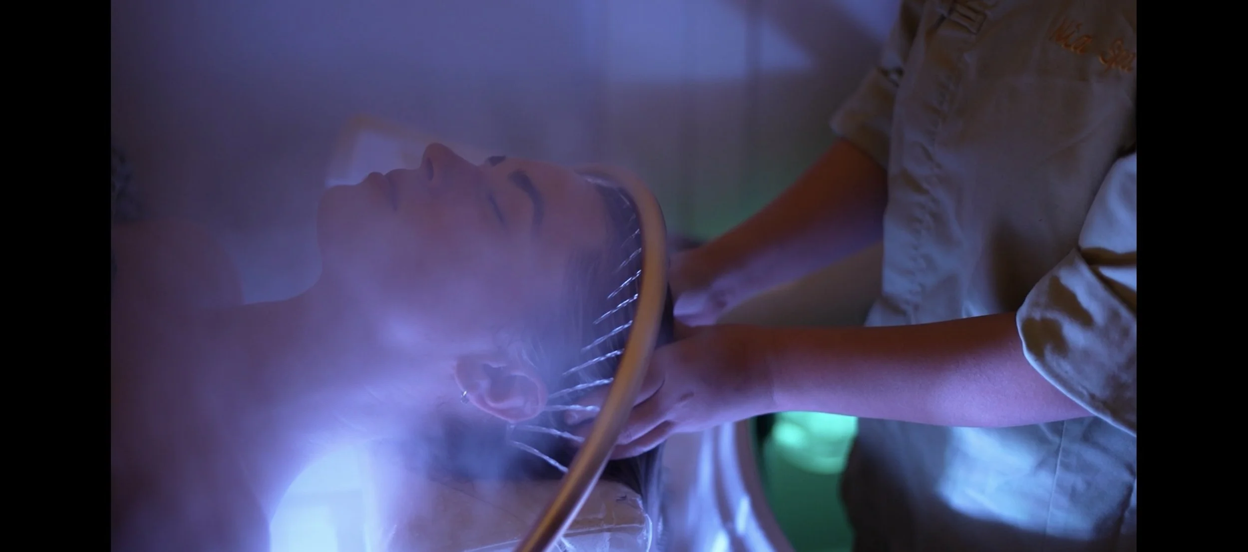 A woman undergoing a medical imaging procedure, lying on her back with head tilted back, while a healthcare professional supports her head under a blue and purple LED light.